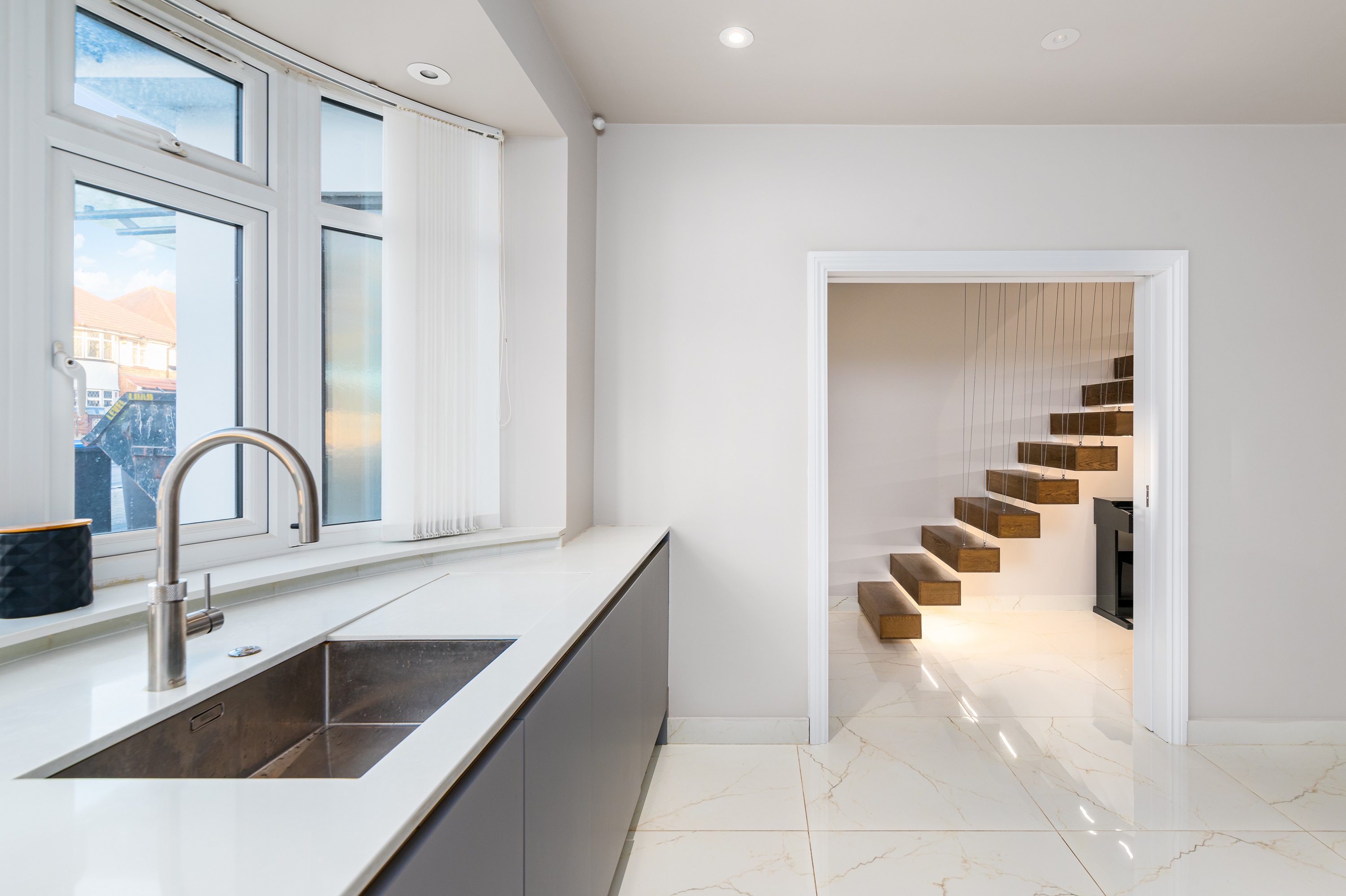 Modern kitchen featuring a sleek sink and countertop, with large windows allowing natural light. A doorway leads to a stylish staircase with wooden steps, showcasing contemporary design elements. An Arch KBB team member is present, demonstrating the space's functionality.