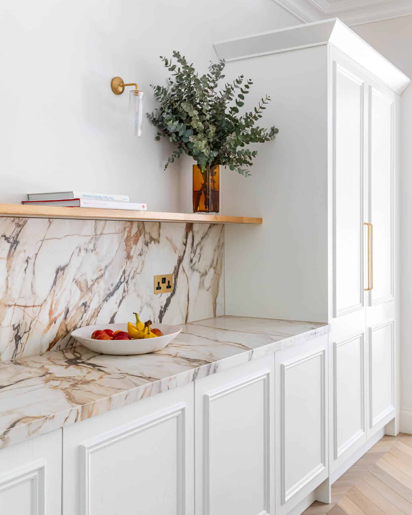 Arch KBB team member showcases a stylish kitchen with elegant marble countertops and a decorative wooden shelf. A bowl of fresh fruit sits on the countertop, complemented by a vase of greenery, creating a modern and inviting atmosphere.