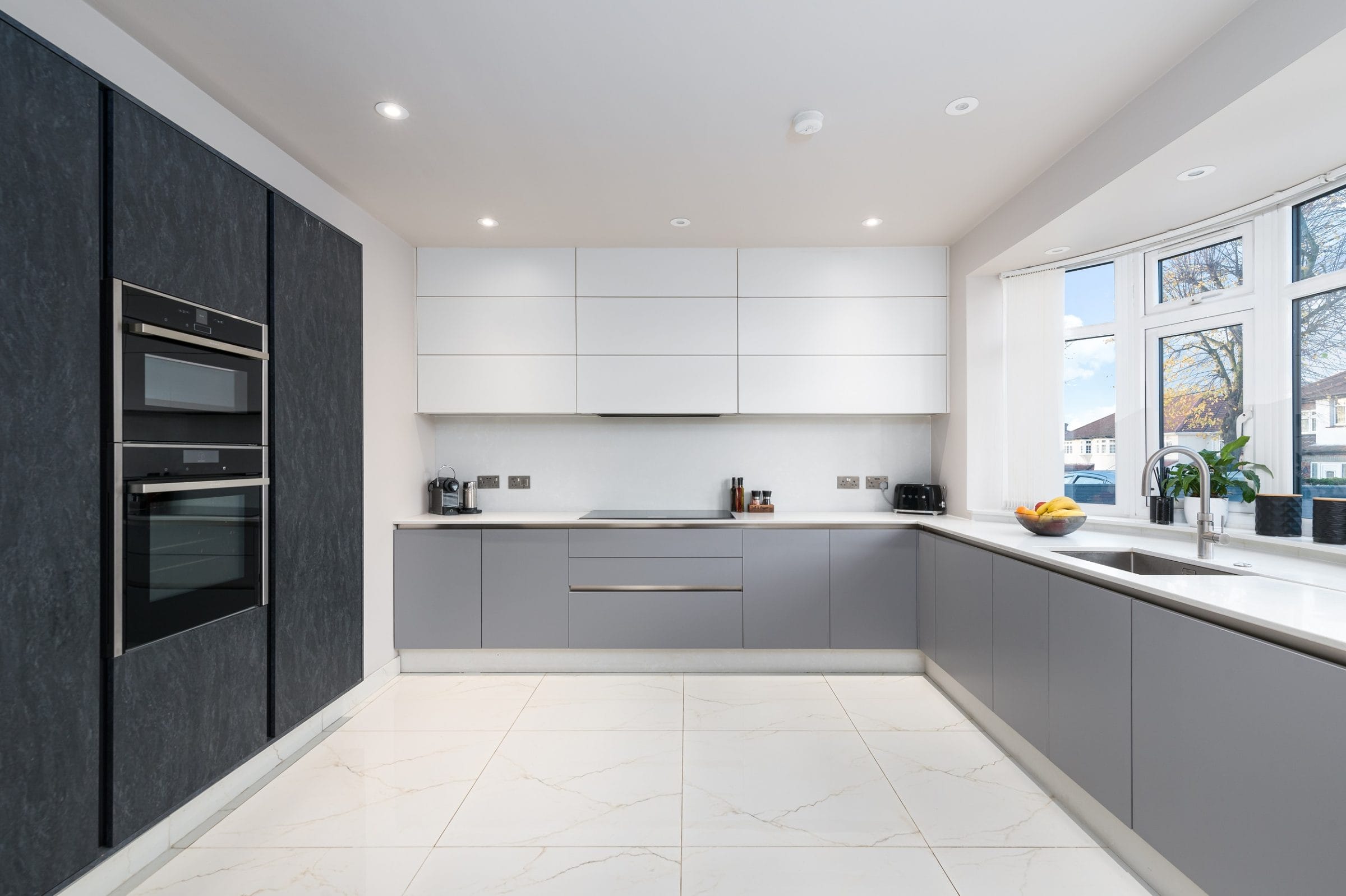 Modern kitchen design featuring sleek grey and white cabinetry, integrated appliances, and a spacious layout. Brightly lit with natural light from the window, showcasing a clean and contemporary aesthetic. An Arch KBB team member is present in the kitchen, enhancing the welcoming atmosphere.
