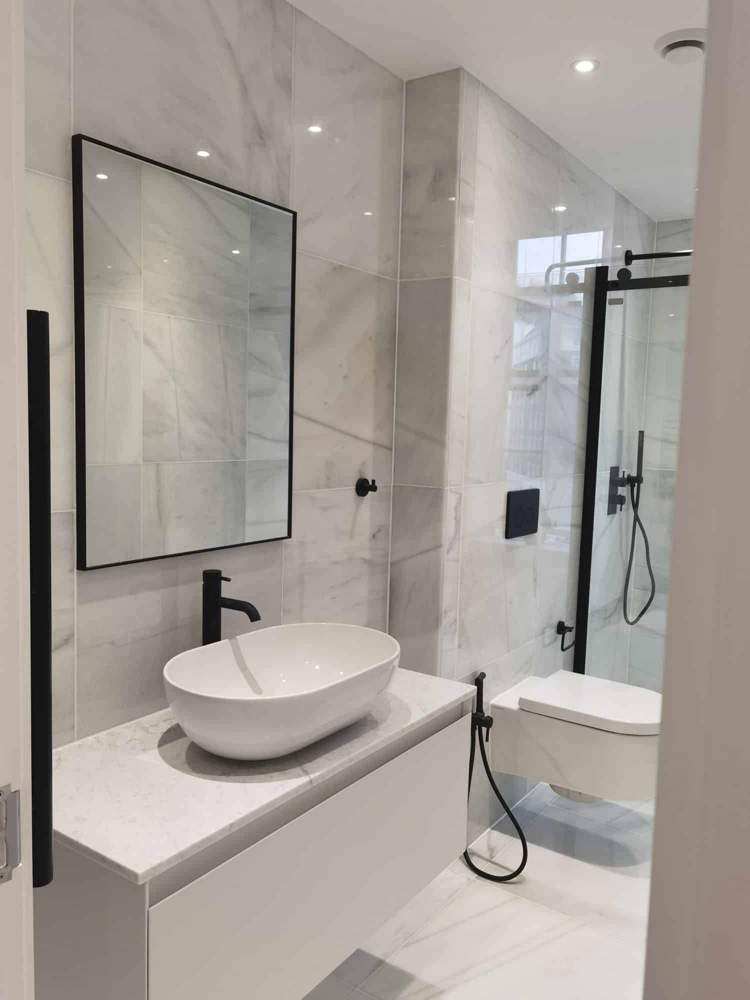 Modern bathroom featuring a sleek oval basin, minimalist black tap, and a frameless glass shower. The walls are adorned with stylish marble tiles, complemented by a large rectangular mirror and a wall-mounted toilet, creating a contemporary and elegant space. An Arch KBB team member is present, showcasing the design.