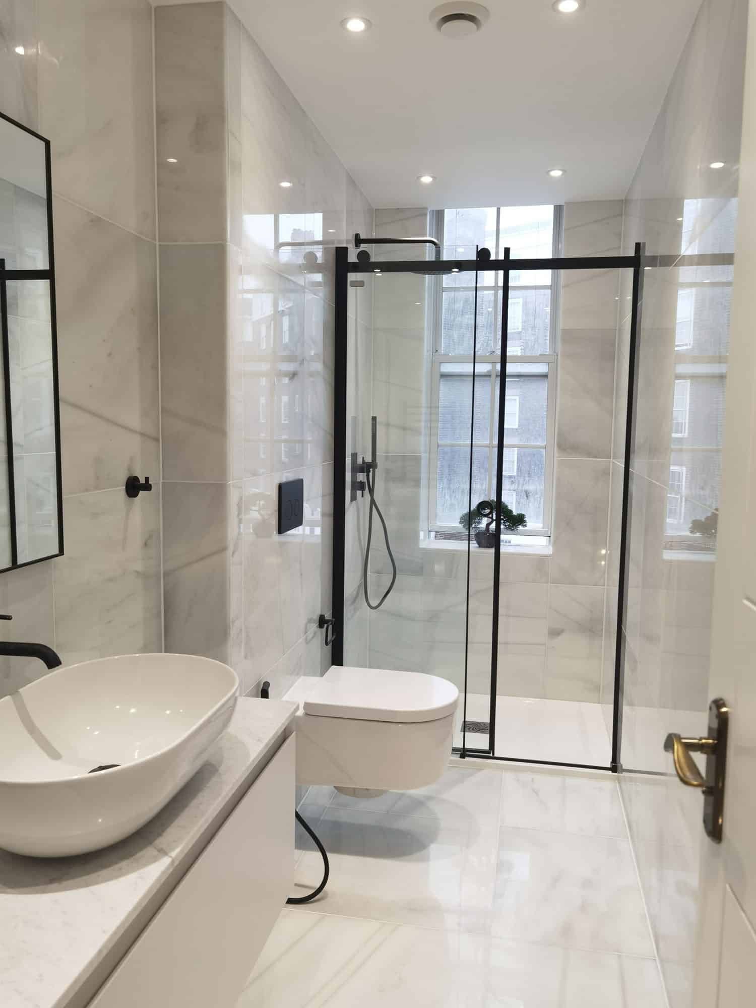 Modern bathroom featuring a sleek design with marble walls, a glass shower enclosure, a wall-mounted toilet, and a stylish basin. An Arch KBB team member is present, enhancing the aesthetic appeal of the space.