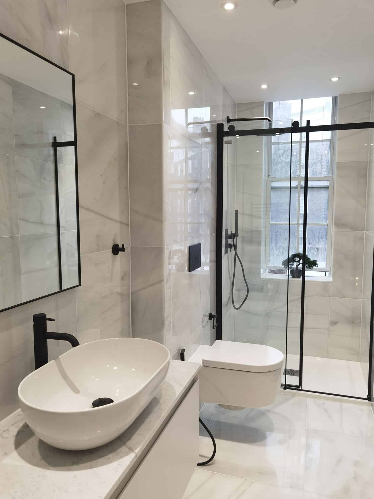 Modern bathroom featuring a sleek design with marble walls, a freestanding basin, a minimalist toilet, and a glass shower enclosure. Natural light streams in through a window, enhancing the contemporary aesthetic. An Arch KBB team member is present, showcasing expert design and installation.