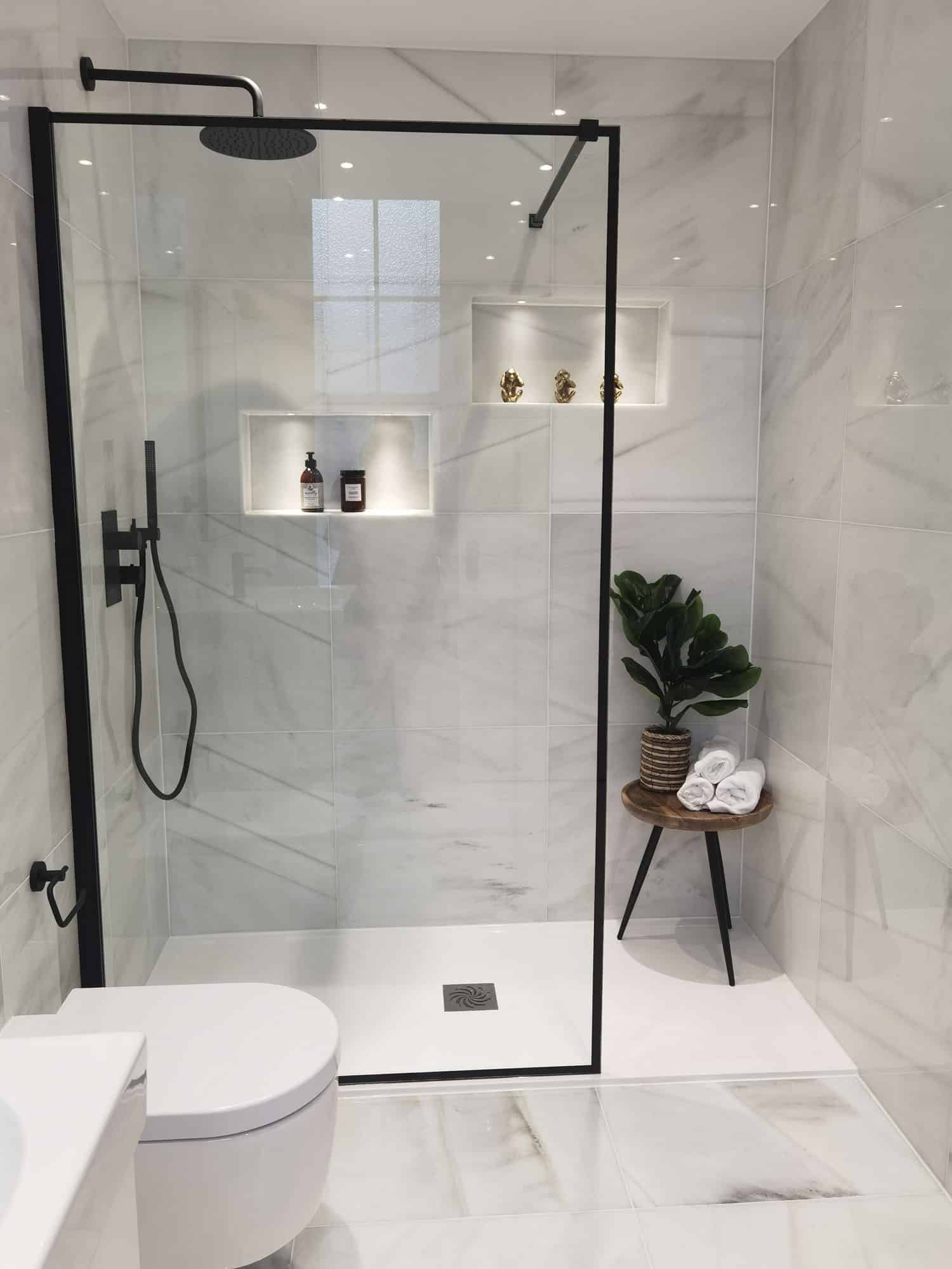 Modern bathroom shower featuring sleek glass enclosure, marble tiles, built-in shelves with decorative items, and a stylish stool with towels and a plant, designed by an Arch KBB team member.