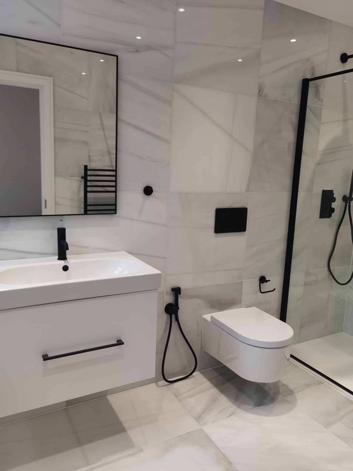 Modern bathroom interior featuring a sleek white vanity with a black faucet, a wall-mounted mirror, and minimalist design elements. The space includes a contemporary toilet and a glass shower enclosure with black fittings, all set against elegant marble tiles. An Arch KBB team member is present, showcasing the stylish and functional design.