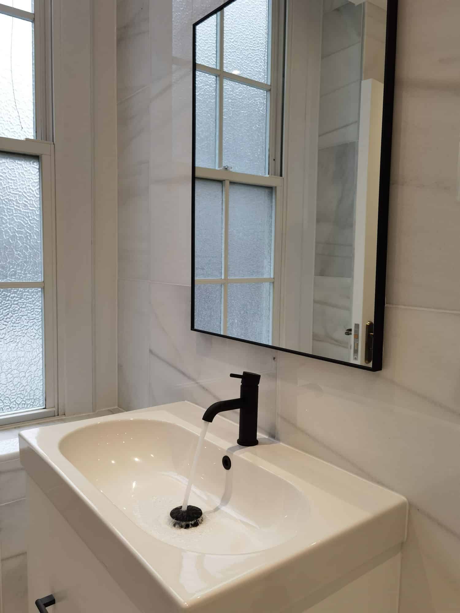 Modern bathroom design featuring a sleek black tap and a white basin, with a large mirror reflecting natural light from a window. The walls are adorned with elegant marble, creating a sophisticated and airy atmosphere. An Arch KBB team member is present, showcasing the stylish installation.