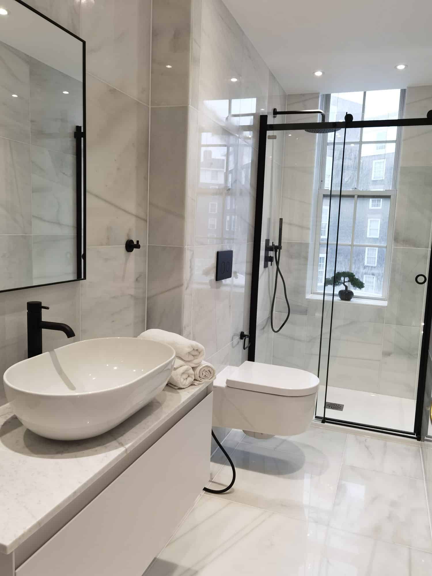 Modern bathroom featuring a stylish freestanding basin, neatly arranged towels, and a sleek shower enclosure. The space is adorned with elegant marble tiles and black fixtures, creating a contemporary aesthetic. An Arch KBB team member is present, showcasing the design's functionality and appeal.