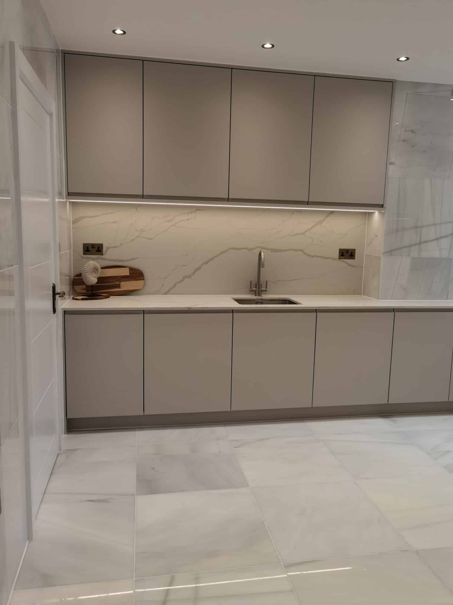 Modern kitchen design featuring sleek grey cabinetry, a minimalist sink area, and stylish lighting. An Arch KBB team member is present, showcasing the elegant marble backsplash and a wooden serving board on the countertop.