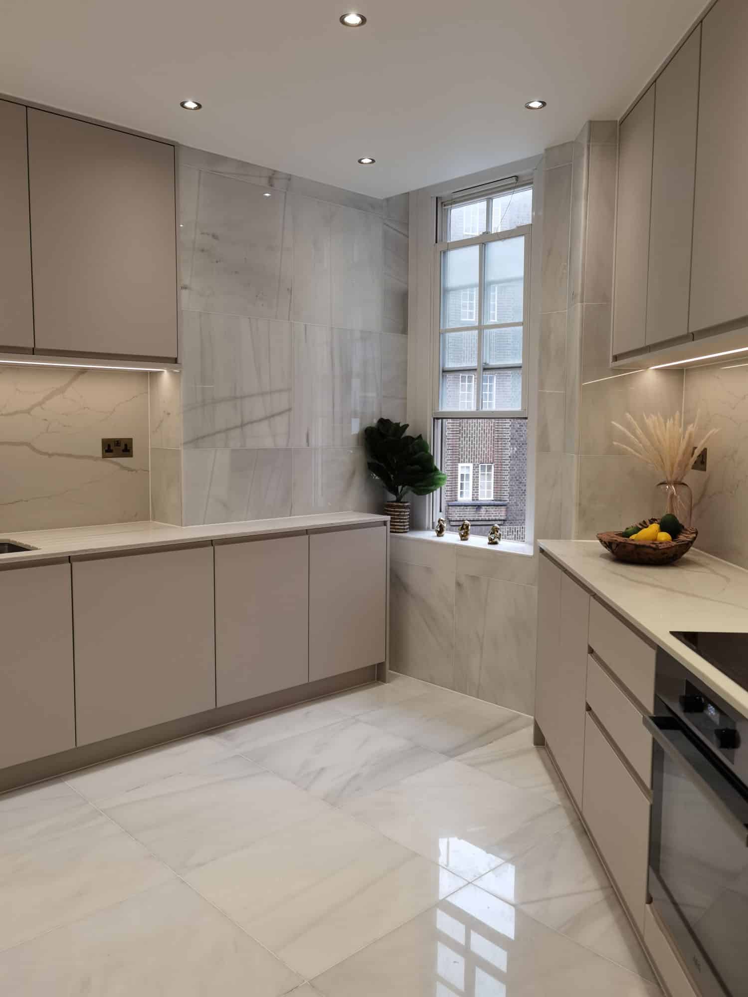Modern kitchen design featuring sleek cabinetry, marble walls, and ambient lighting. The space includes a window with decorative plants and a fruit bowl, creating a bright and inviting atmosphere. An Arch KBB team member is present, engaged in the space.