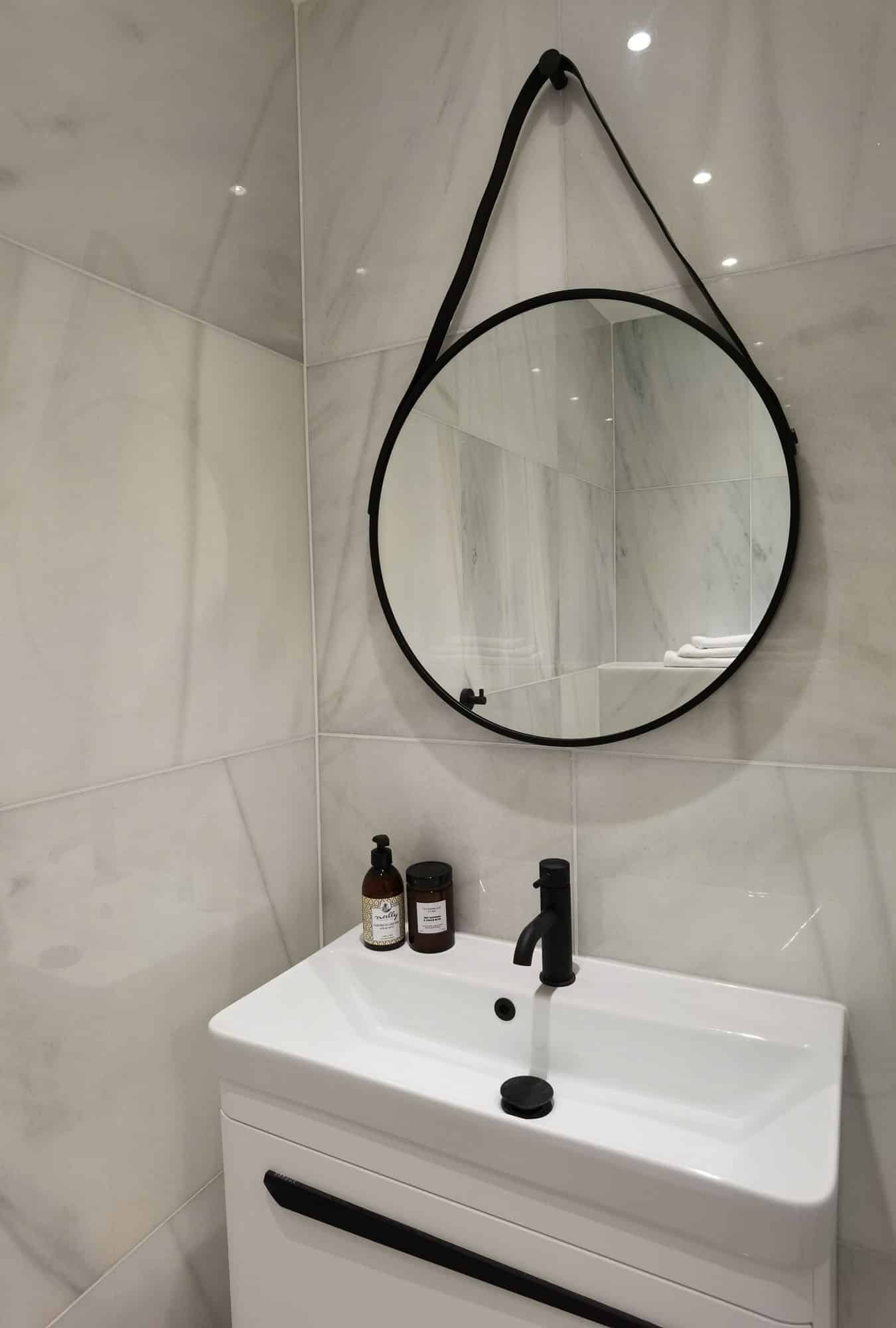 Modern bathroom interior featuring a circular mirror above a sleek sink, with stylish toiletries displayed nearby. The marble-effect tiles add a touch of elegance to the space. An Arch KBB team member is present, contributing to the design aesthetic.