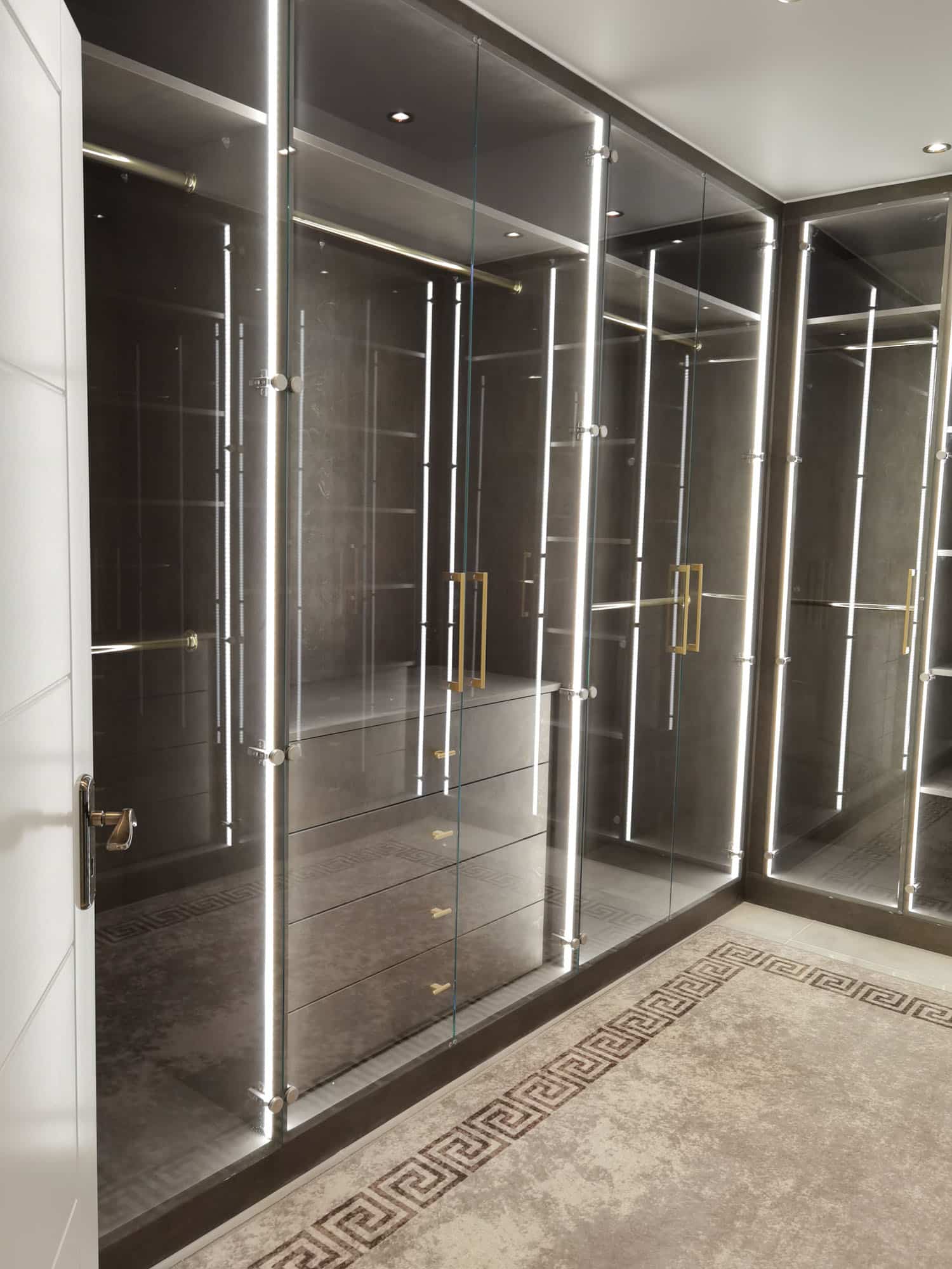 Interior view of a modern wardrobe featuring sleek glass doors, illuminated by stylish LED lighting, showcasing a contemporary design with elegant gold handles. Arch KBB team member can be seen in the background, enhancing the luxurious setting.