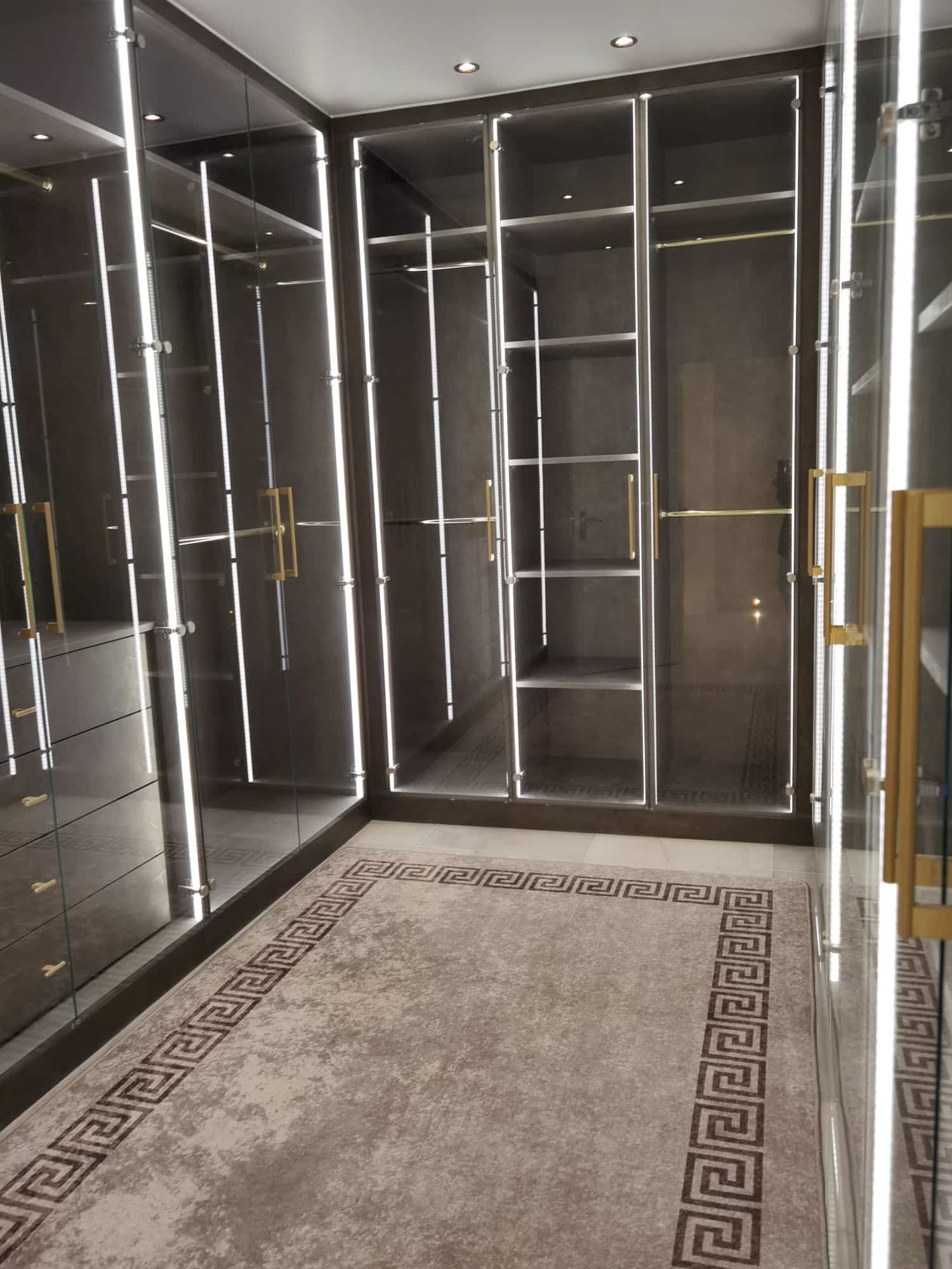 A modern, stylish wardrobe space featuring glass-fronted storage units with integrated lighting. The design includes shelves and drawers, complemented by a decorative rug with a geometric pattern. An Arch KBB team member is present, showcasing the functional and aesthetic appeal of the storage solution.