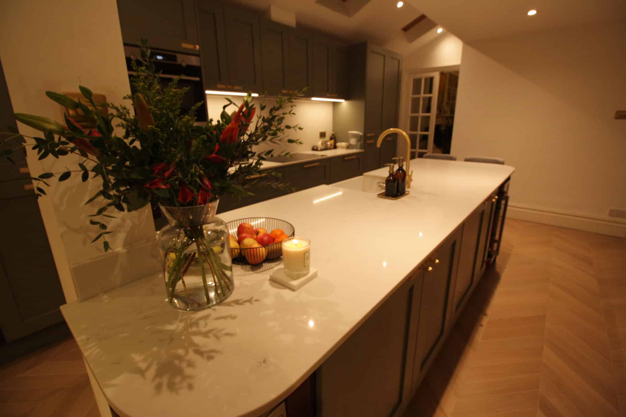 Modern kitchen interior featuring a stylish island with a marble countertop, decorative flowers, fruit bowl, and candle. The space is illuminated by warm lighting, showcasing sleek cabinetry and a contemporary design. An Arch KBB team member can be seen in the background, enhancing the inviting atmosphere.