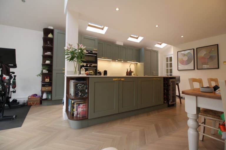 Modern kitchen design featuring a stylish green cabinetry, well-lit by skylights, with an open layout. The space includes a dining area and is complemented by decorative artwork on the walls. An Arch KBB team member is present, showcasing the inviting atmosphere of the kitchen.