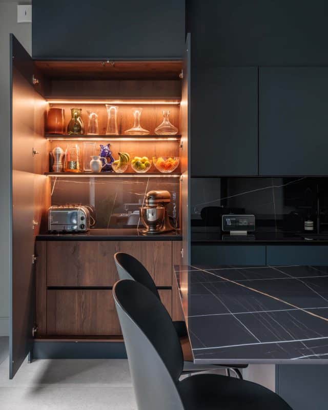 Modern kitchen featuring a stylish cabinet with illuminated glass shelves displaying various kitchenware, including glassware and a stand mixer. The sleek countertop complements the contemporary design, with an Arch KBB team member visible in the background.