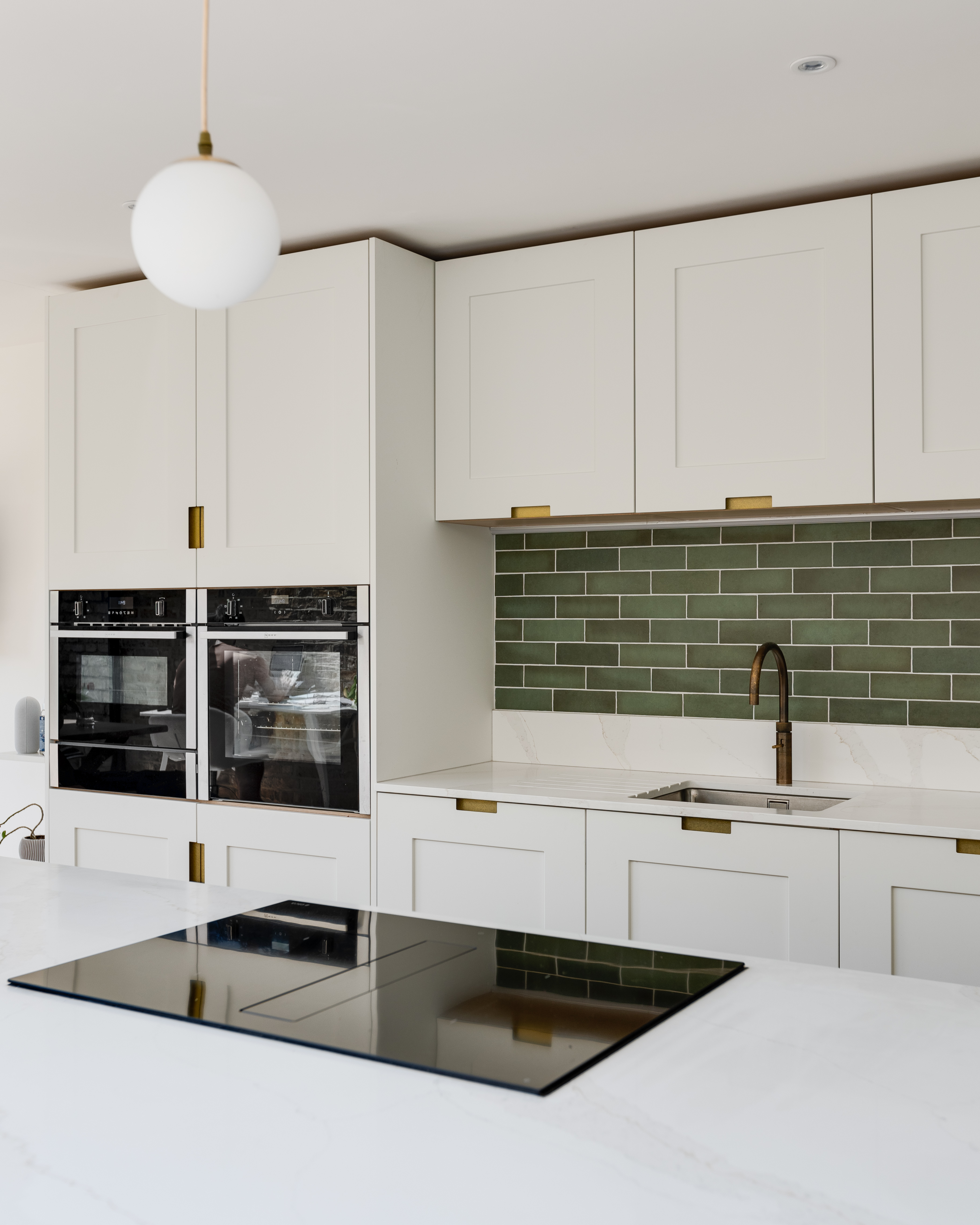 Modern kitchen design featuring sleek white cabinetry, integrated double ovens, and a stylish green backsplash. The space includes a minimalist sink with a brass tap and a contemporary induction cooktop, showcasing a blend of functionality and aesthetic appeal, designed by an Arch KBB team member.