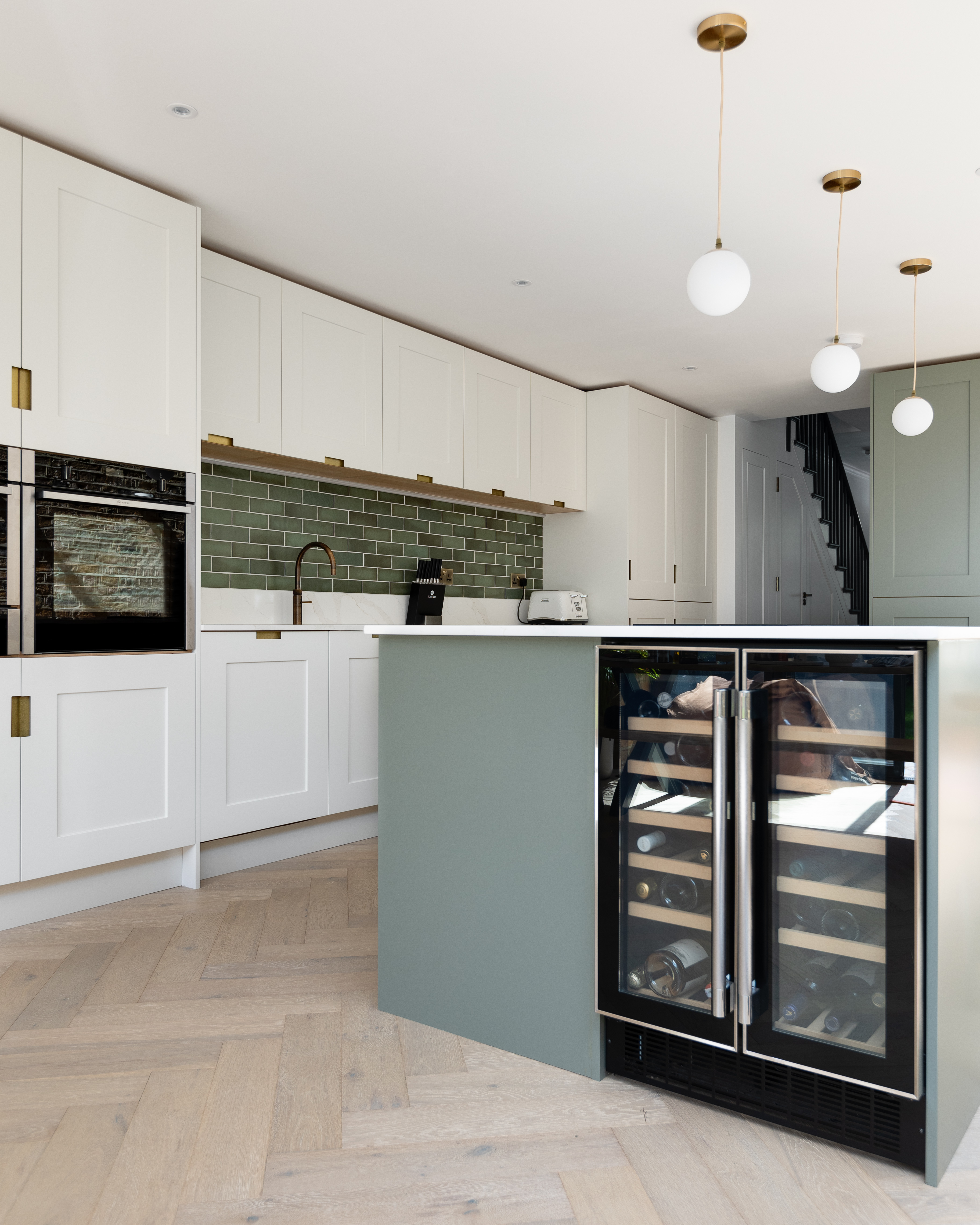Modern kitchen interior featuring sleek white cabinetry, a stylish green tiled backsplash, and a central island with a wine cooler. An Arch KBB team member is present, enhancing the welcoming atmosphere of this contemporary space.