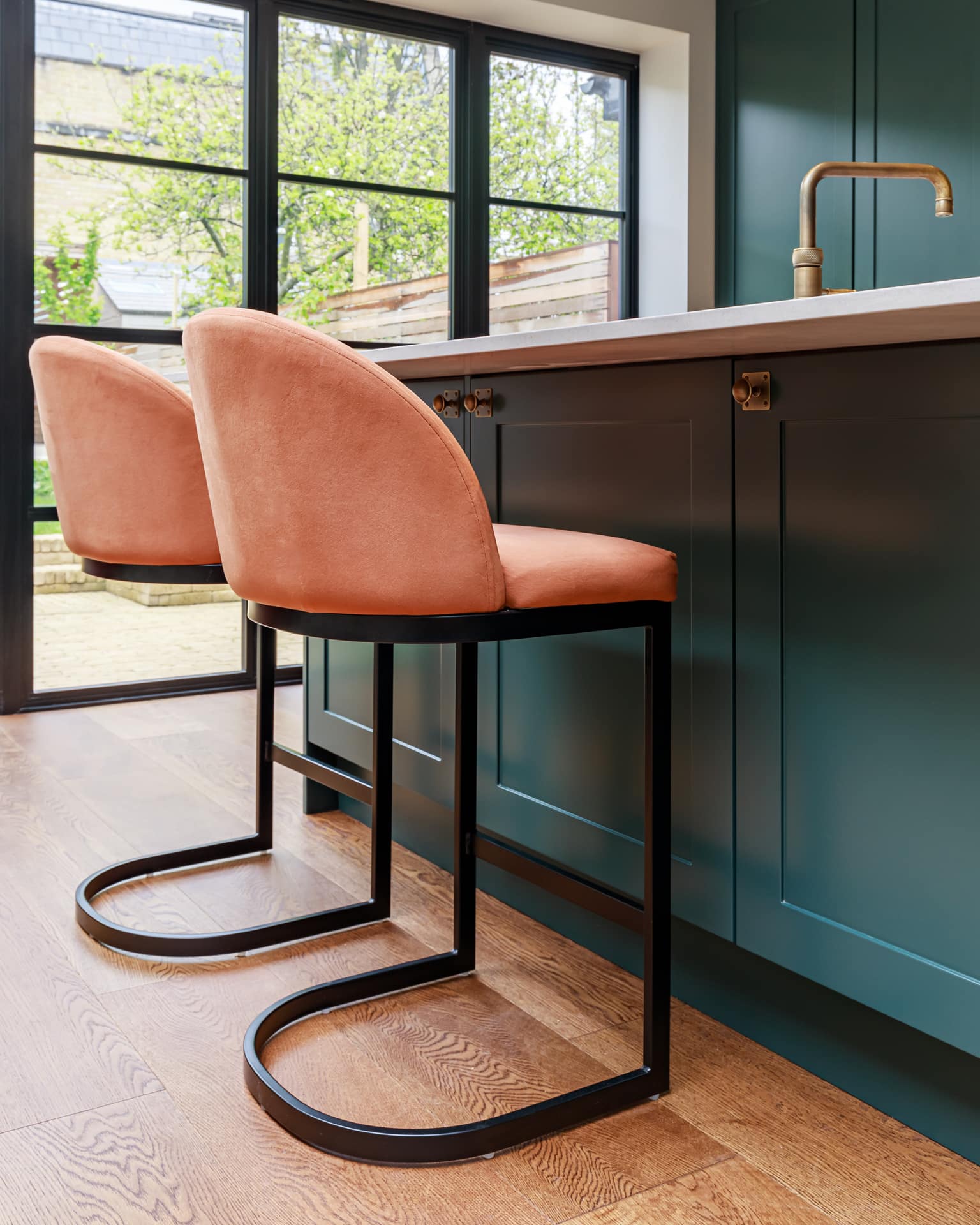 Interior view of a modern kitchen featuring two stylish peach upholstered bar stools with black metal frames positioned at a sleek countertop, complemented by dark cabinetry and large windows allowing natural light to fill the space. An Arch KBB team member is present, engaged in the design process.