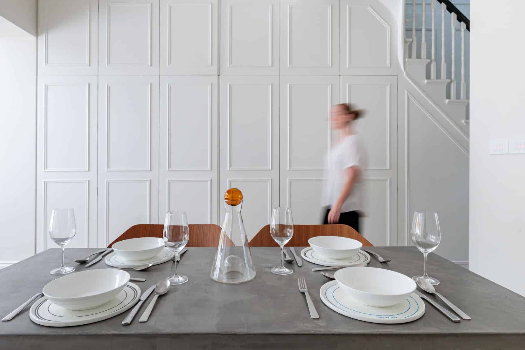 Dining table set with white plates, glassware, and cutlery, featuring a decorative carafe with an orange accent. An Arch KBB team member is seen moving in the background, against a stylish white paneled wall. The scene showcases a modern and minimalist dining setting.