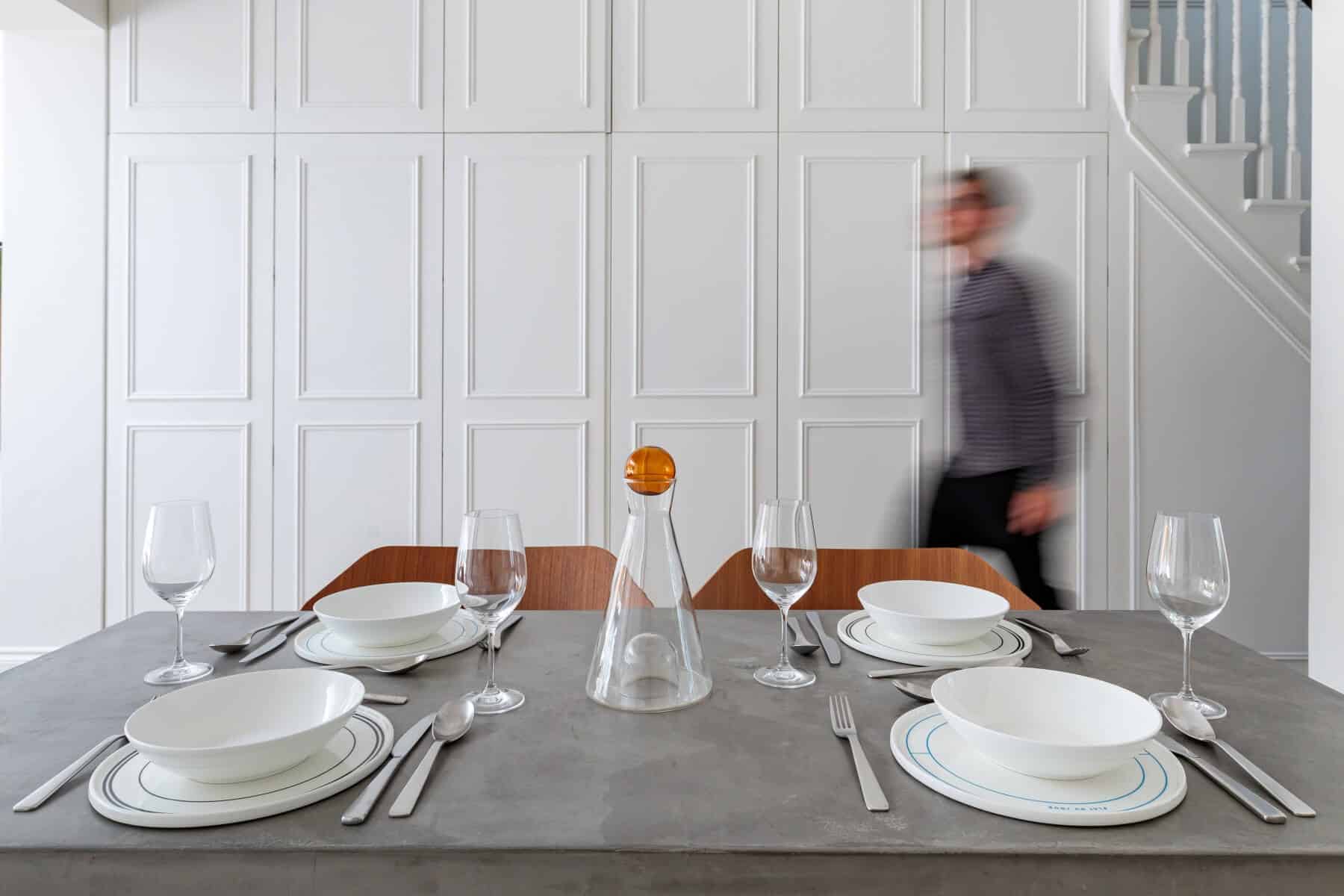 A modern dining table set with white plates, glasses, and cutlery, featuring a decorative glass carafe with an orange top. An Arch KBB team member is blurred in motion in the background, adding a dynamic element to the contemporary interior design. The wall behind showcases elegant paneling, enhancing the sophisticated atmosphere of the space.