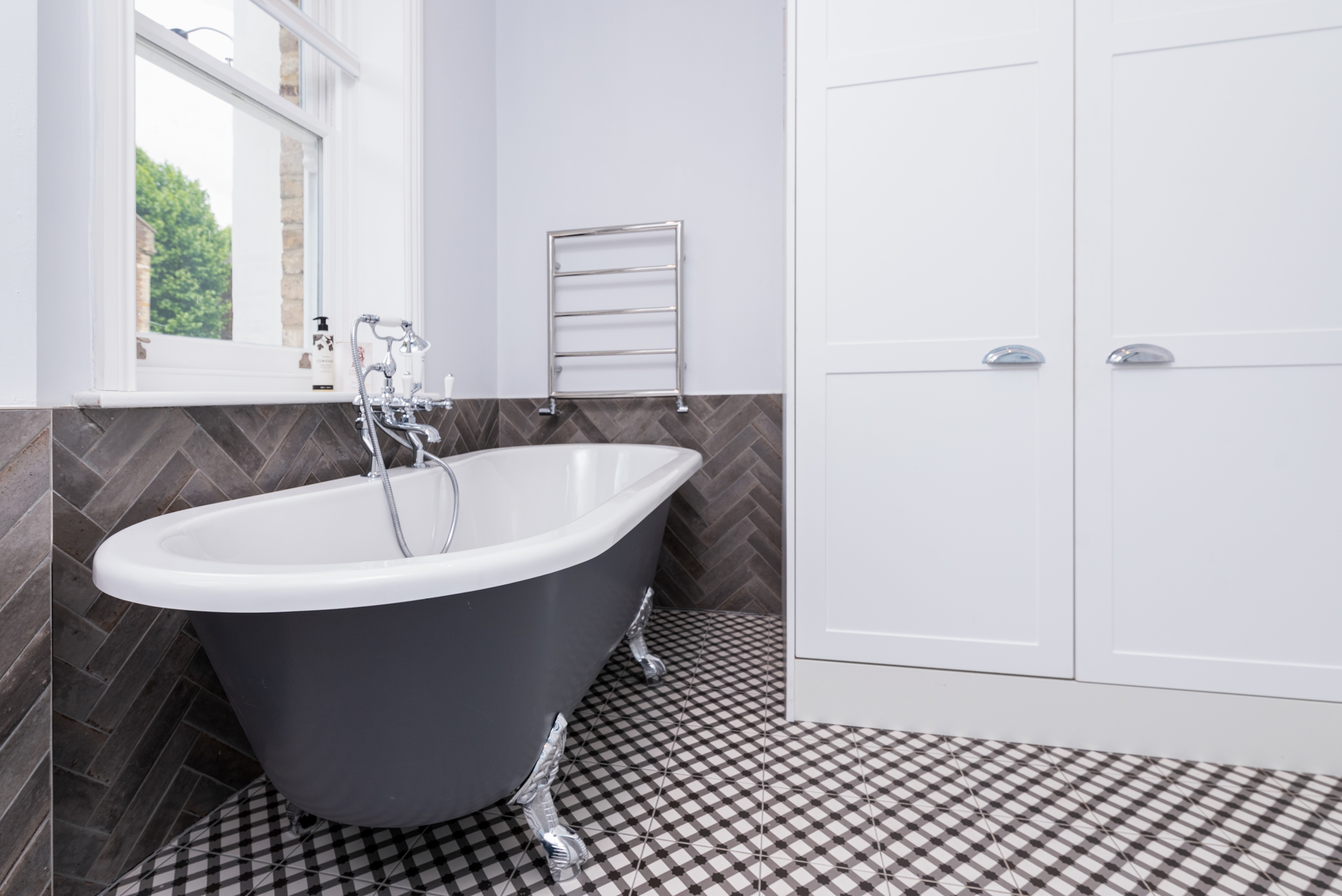A stylish bathroom featuring a freestanding bath with a grey exterior, chrome fixtures, and a modern towel rail. The walls are painted in a soft shade, complementing the herringbone patterned floor tiles. An Arch KBB team member is present, showcasing the elegant design and layout.