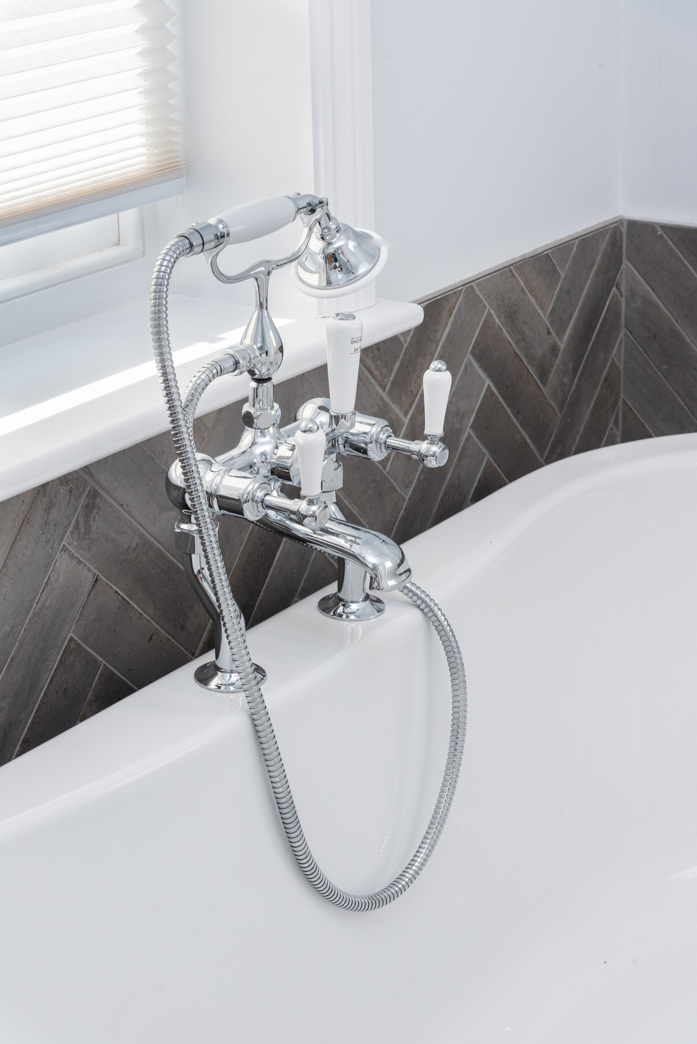 Chrome bath tap with a handheld showerhead, featuring white ceramic handles, installed on a modern bathtub against a herringbone tiled wall. Arch KBB team member present, showcasing stylish bathroom fittings.