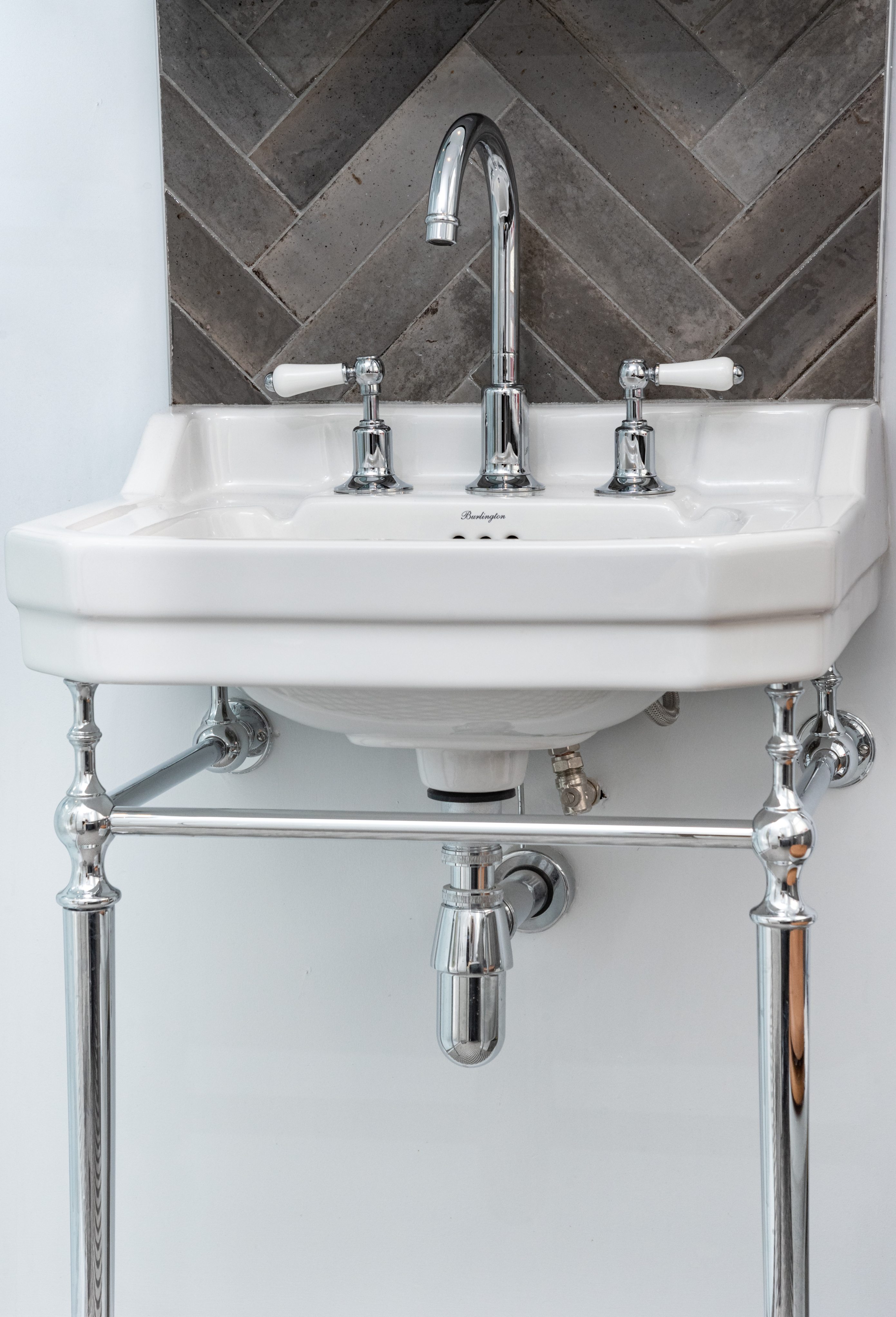 Close-up view of a stylish white sink with chrome taps and elegant legs, set against a textured tiled wall, showcasing a blend of modern and classic design elements. An Arch KBB team member is present, highlighting the attention to detail in kitchen and bathroom installations.
