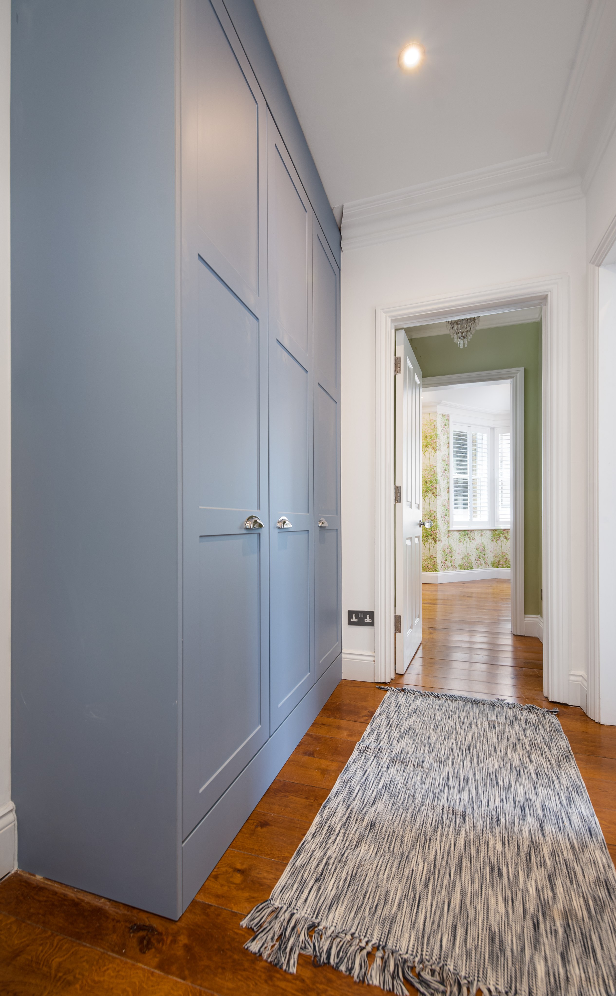 Interior view of a hallway featuring stylish blue fitted wardrobes, wooden flooring, and an open doorway leading to a well-lit room with patterned walls. An Arch KBB team member is present, enhancing the design consultation experience.