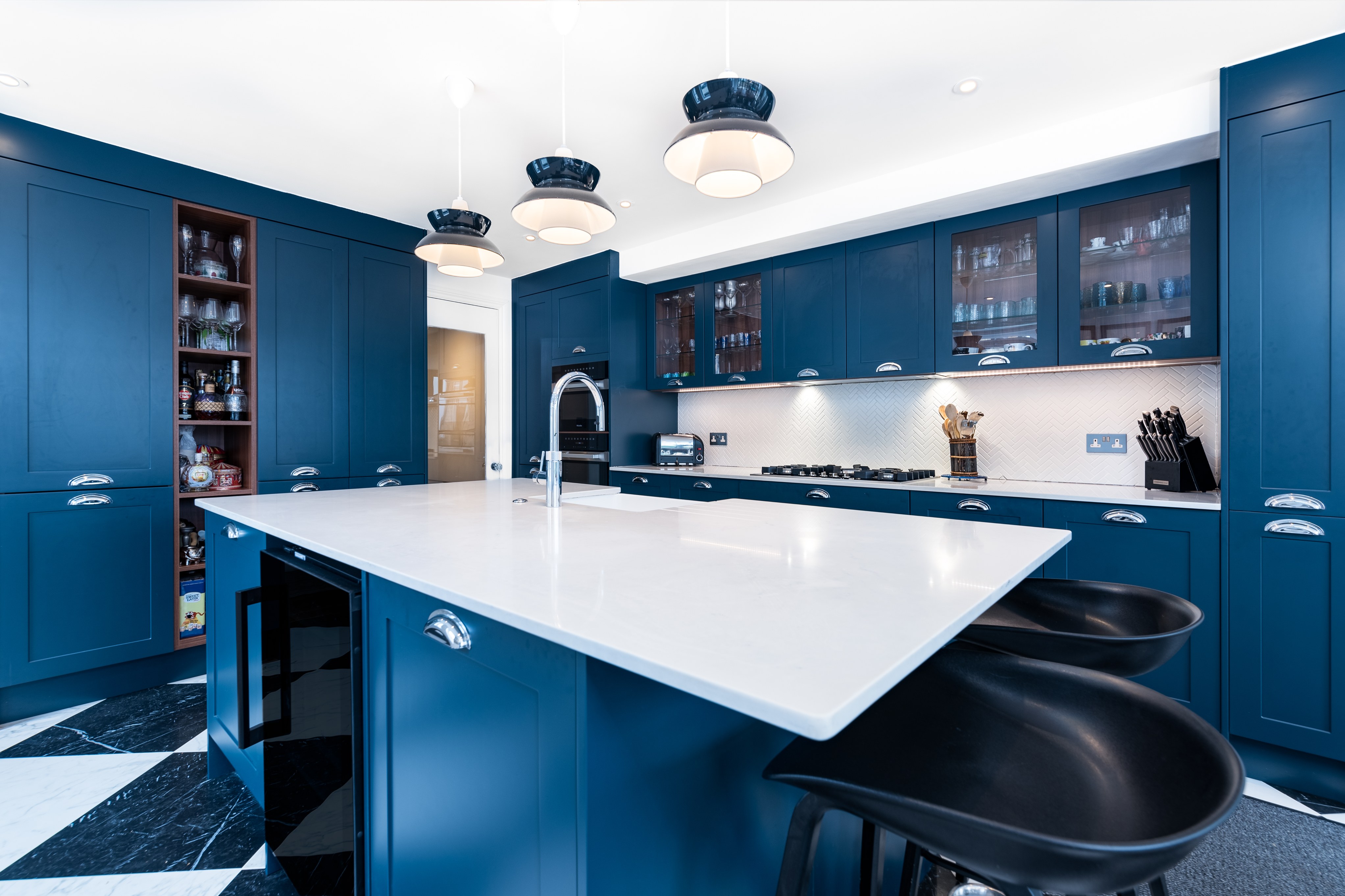 Modern kitchen design featuring deep blue cabinetry, a large white island with seating, and stylish pendant lighting. The space includes a sleek countertop, integrated appliances, and a well-organised pantry area. An Arch KBB team member is present, showcasing the kitchen's contemporary aesthetic and functionality.