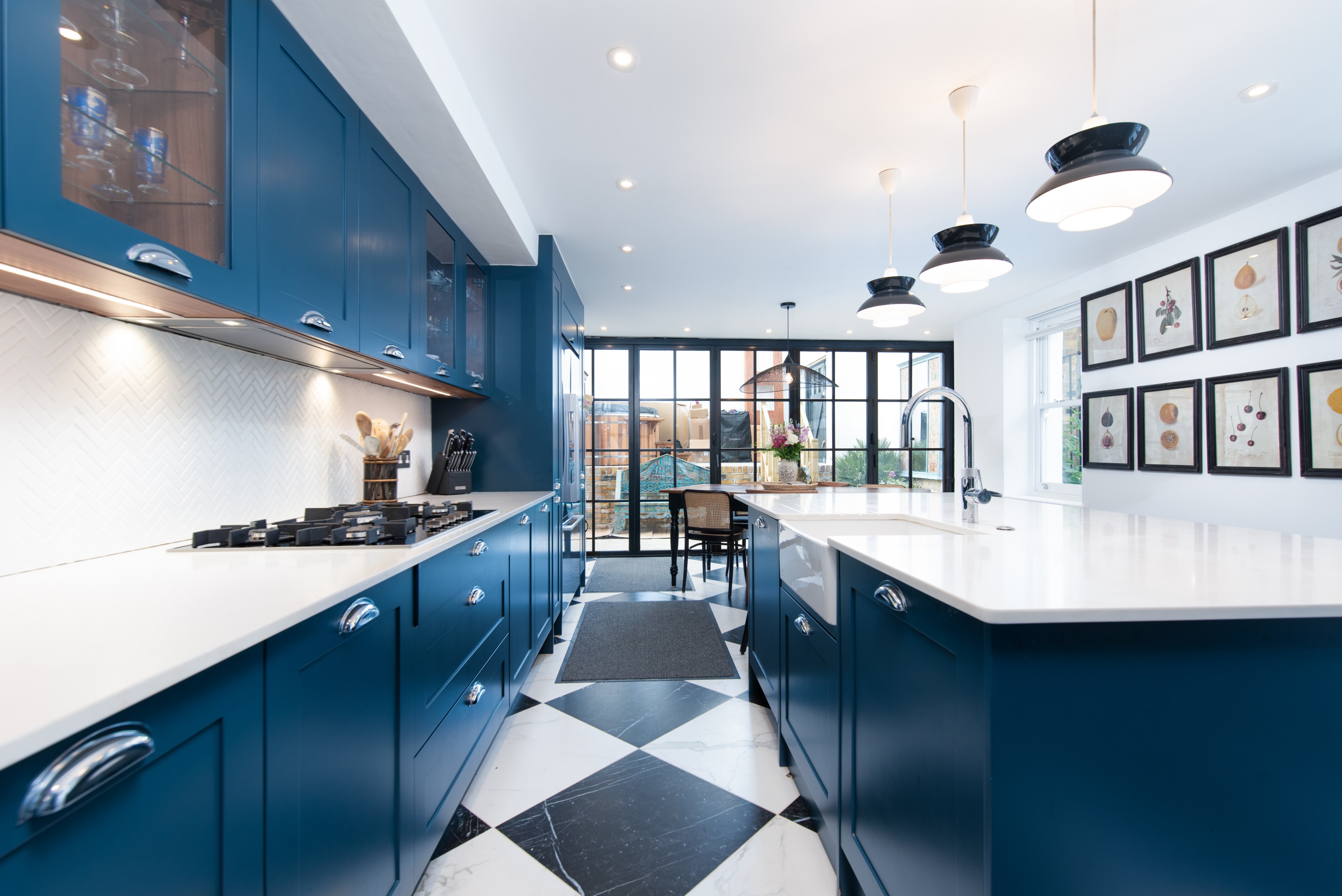 Modern kitchen featuring deep blue cabinetry, white countertops, and a striking black and white tiled floor. Brightly lit space with large windows and outdoor views, complemented by stylish pendant lighting. An Arch KBB team member is present, enhancing the kitchen design experience.