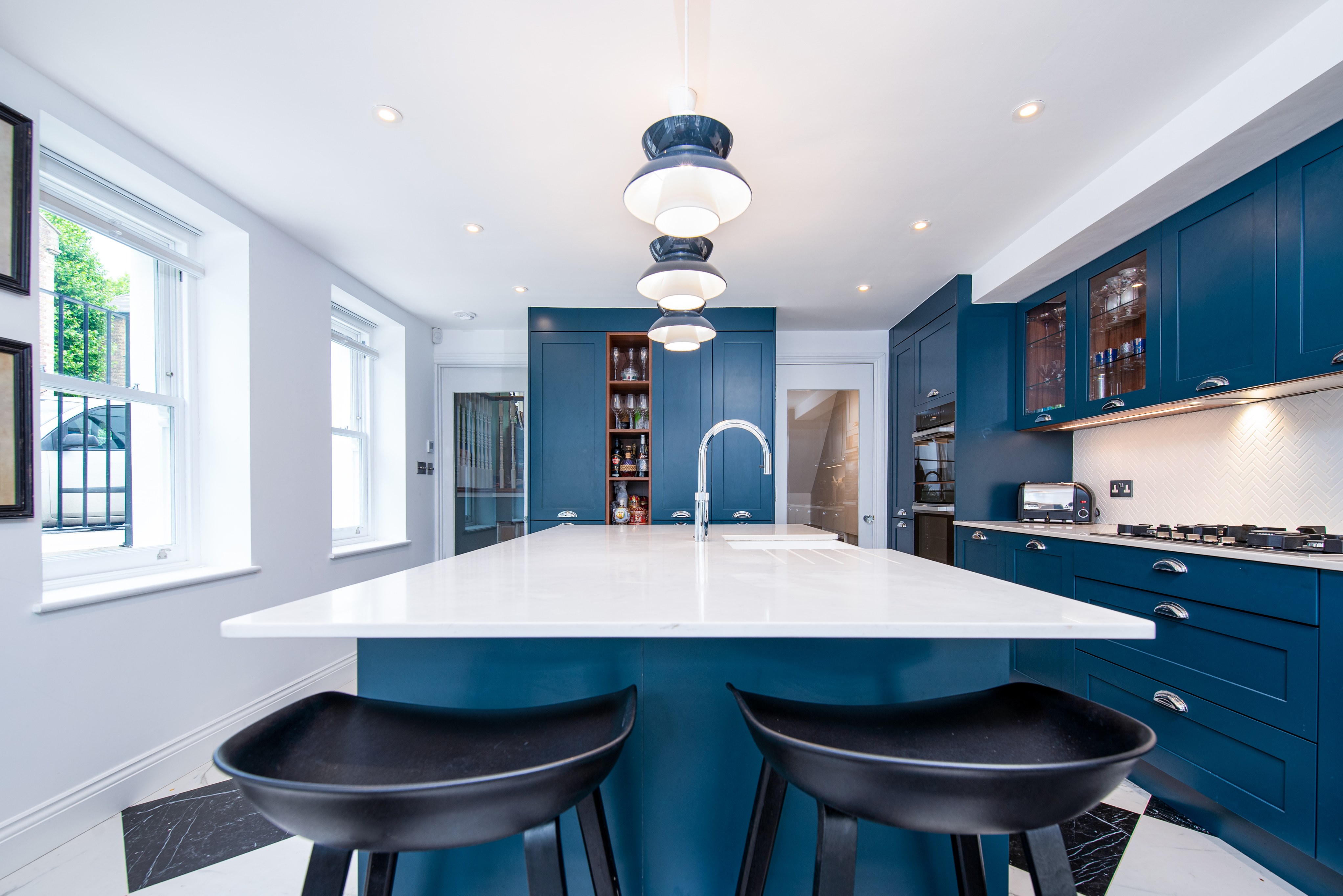 Modern kitchen design featuring a spacious island with a white countertop, blue cabinetry, and stylish pendant lighting. The room is well-lit with natural light from large windows, enhancing the contemporary aesthetic. An Arch KBB team member is present, showcasing the kitchen's functional layout.