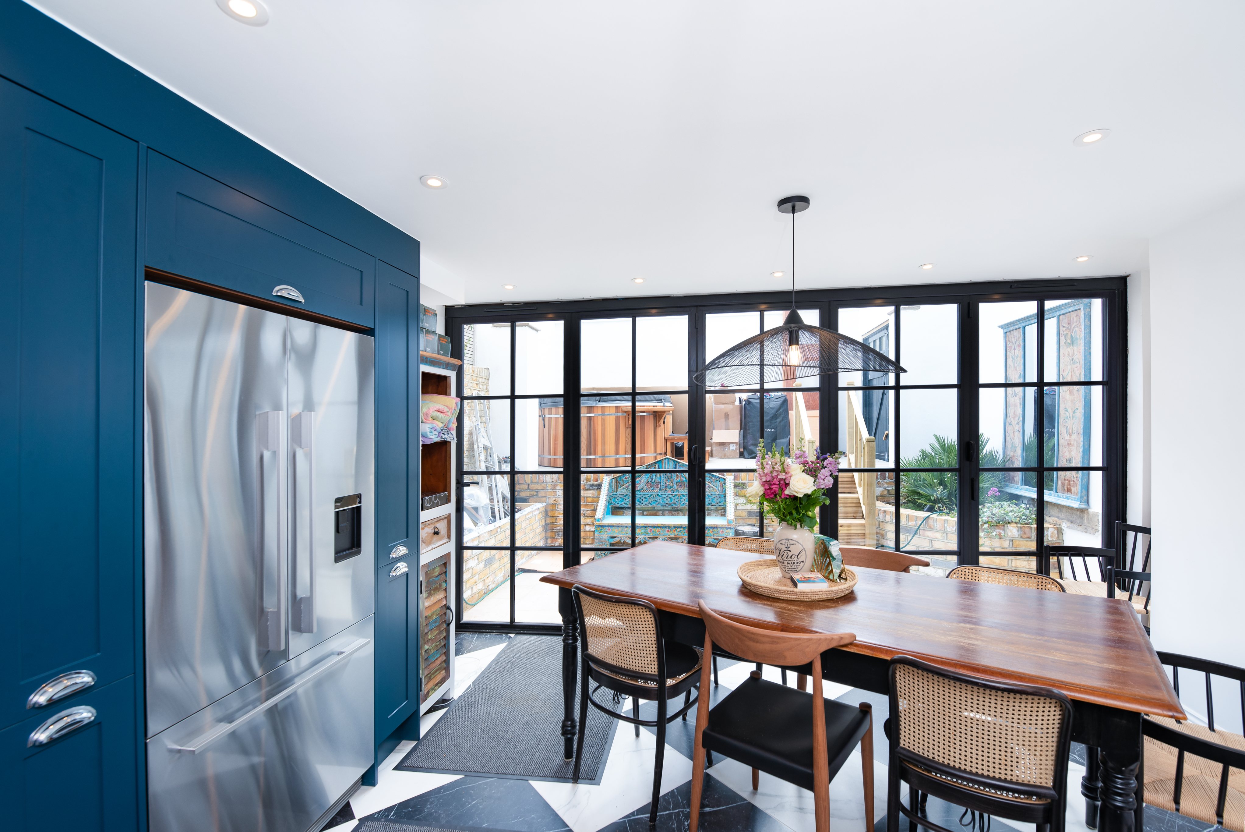 Modern kitchen featuring a stylish dining table and black-framed glass doors, allowing natural light to illuminate the space. An Arch KBB team member is present, and the kitchen is equipped with sleek appliances, including a stainless steel fridge. The decor combines contemporary design with a welcoming atmosphere, highlighted by floral arrangements on the table.