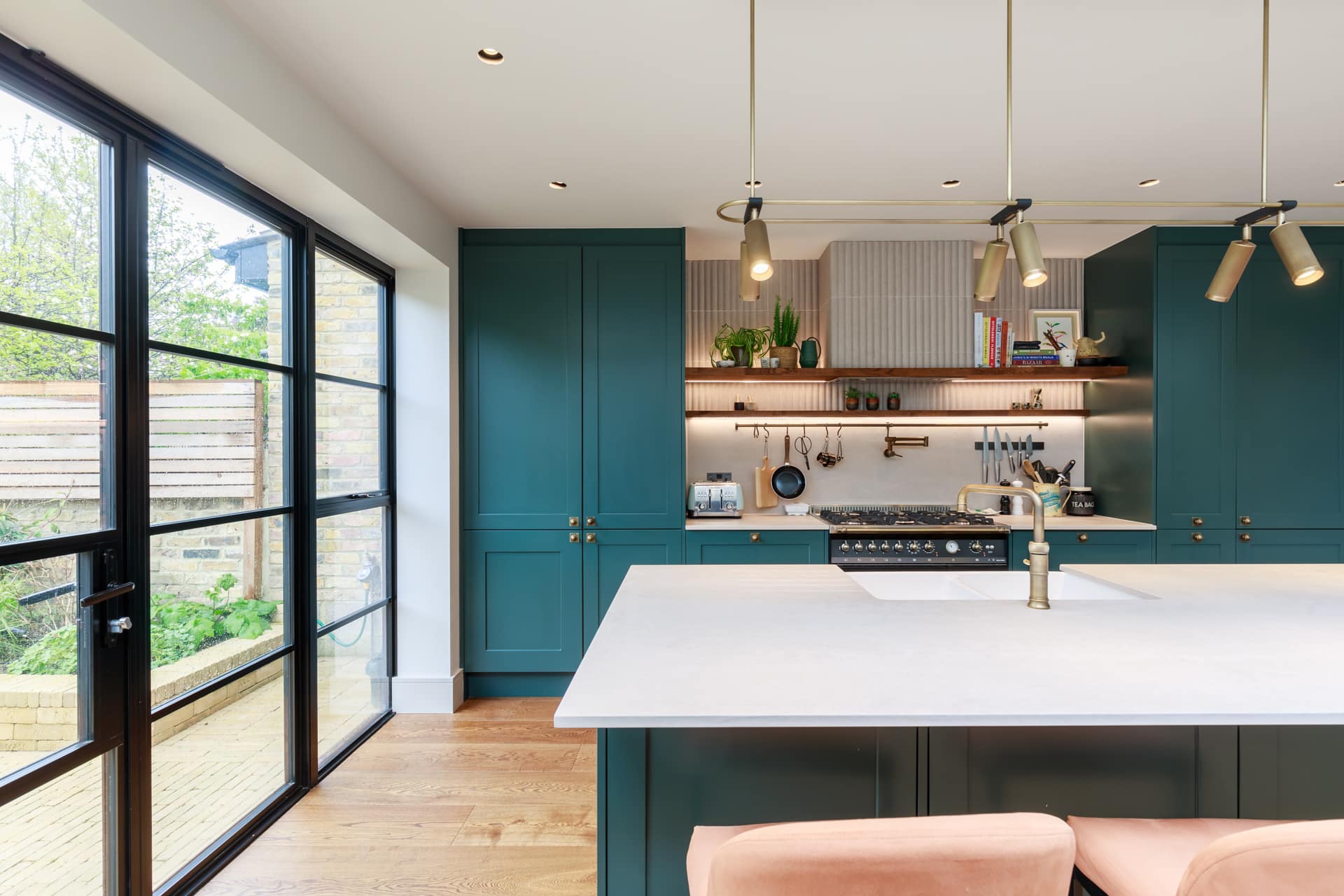 Modern kitchen featuring a spacious island with a white countertop, teal cabinetry, and stylish pendant lights. The design includes open shelving with plants and kitchenware, alongside a large window that offers natural light and a view of the outdoor space. An Arch KBB team member is present, showcasing the contemporary layout and aesthetic.