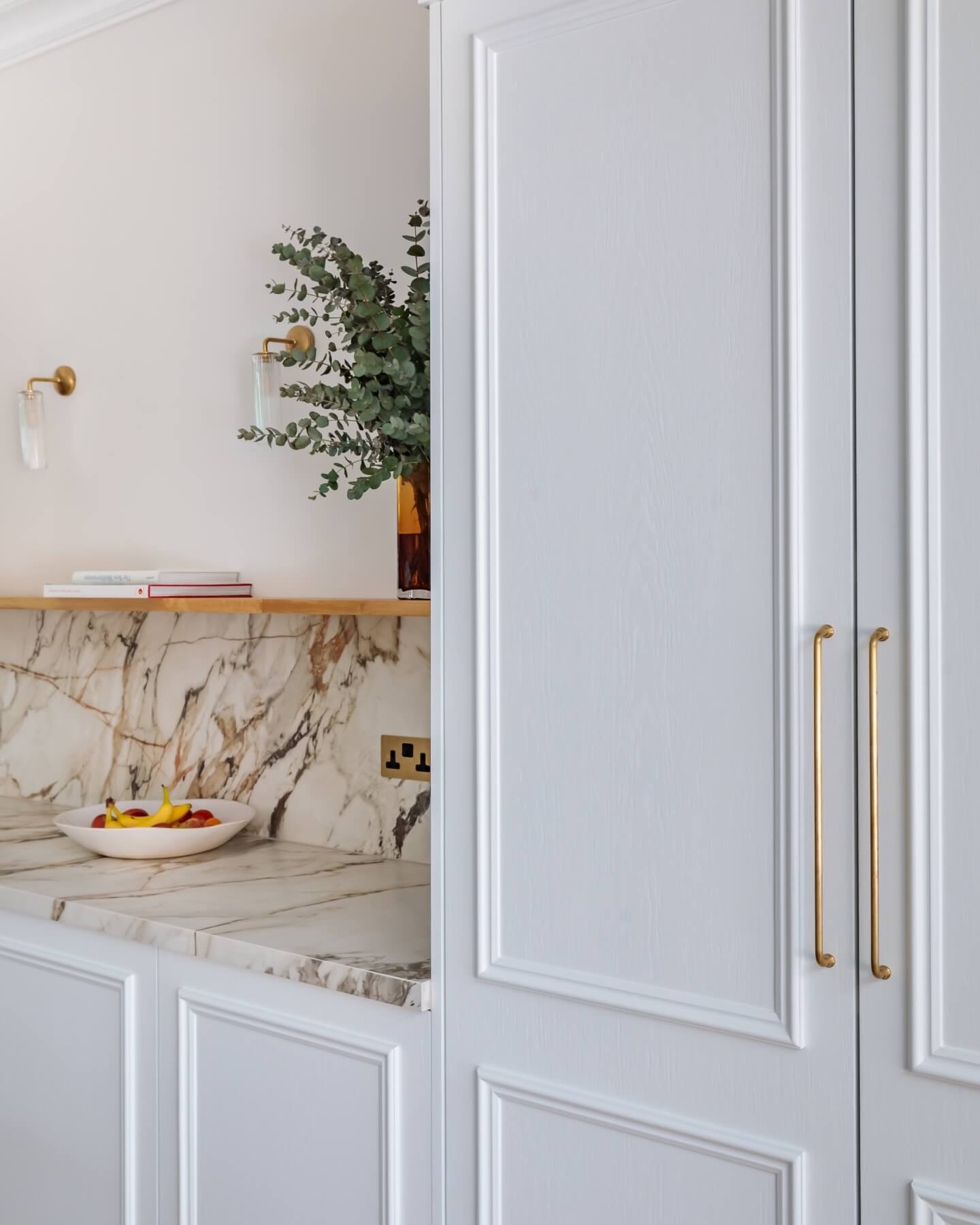 Modern kitchen interior featuring a marble countertop, a bowl of fresh fruit, and decorative greenery. An Arch KBB team member is present, showcasing stylish cabinetry with gold handles and a minimalist design.