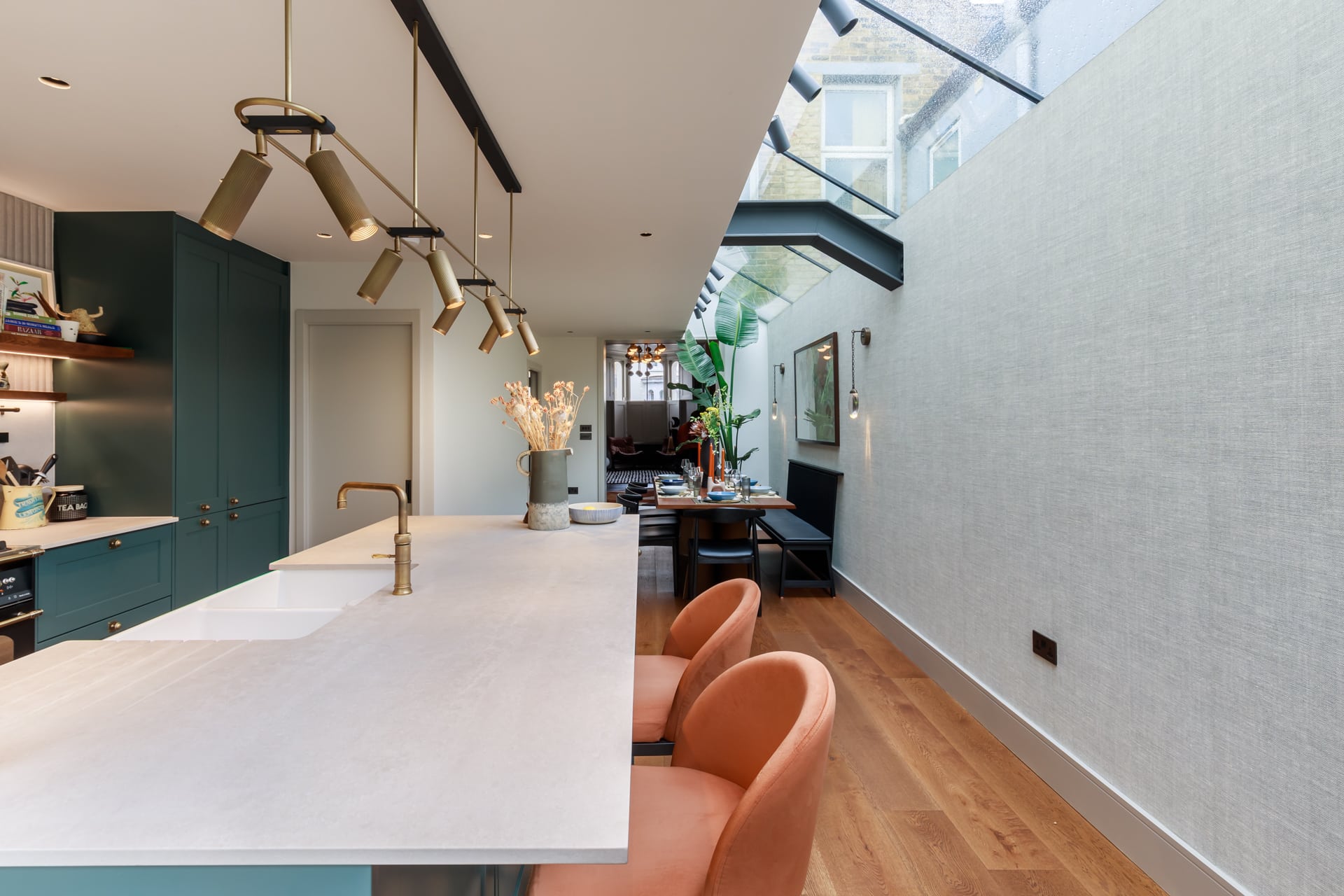 Modern kitchen interior featuring a stylish island with a white countertop, brass fixtures, and pastel pink chairs. The open space is illuminated by natural light through a glass roof, complemented by plants and contemporary decor in the dining area. An Arch KBB team member is present, enhancing the inviting atmosphere.