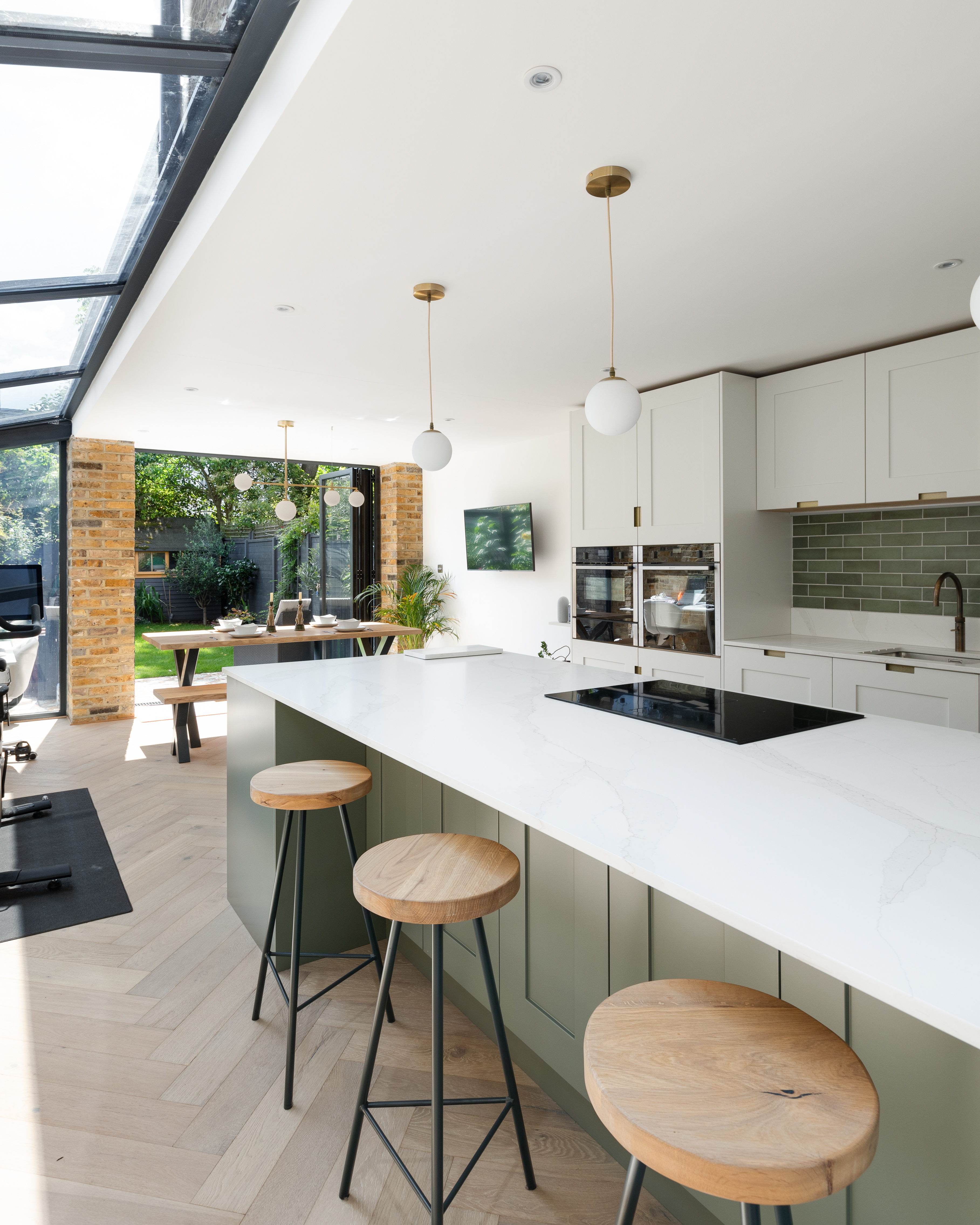 Modern kitchen featuring a spacious island with bar stools, sleek cabinetry, and integrated appliances. Large windows provide natural light and a view of the garden. An Arch KBB team member is present, showcasing the contemporary design and open-plan layout.