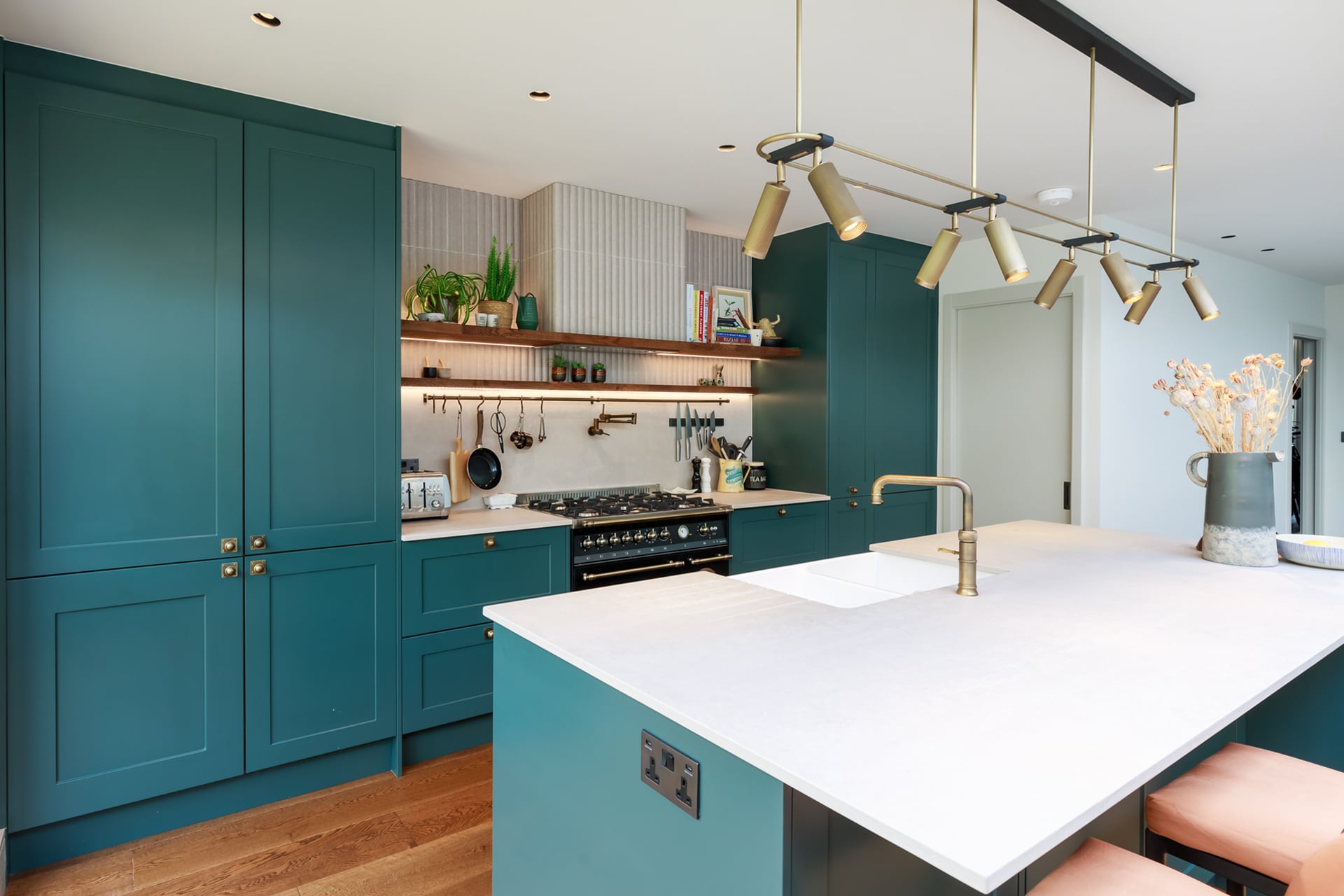 Modern kitchen design featuring teal cabinetry, a spacious island with a white countertop, and stylish pendant lighting. The kitchen is equipped with a gas stove and open shelving displaying plants and kitchenware. An Arch KBB team member is present, highlighting the contemporary aesthetic and functionality of the space.