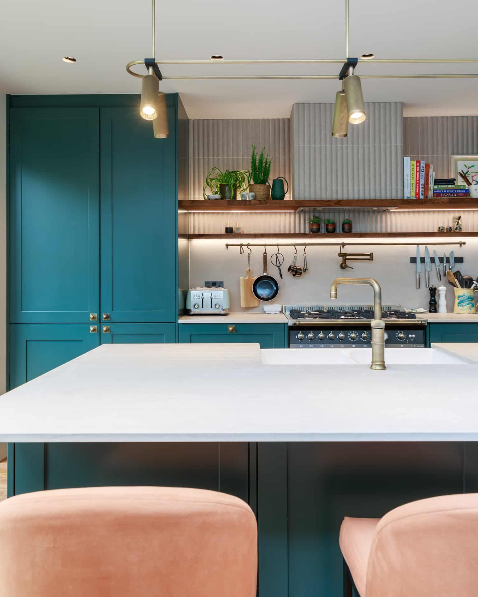Modern kitchen featuring a stylish design with teal cabinetry, a spacious white countertop, and a brass tap. Open shelving displays plants and kitchenware, while a gas range and toaster are visible in the background. An Arch KBB team member is present, showcasing the kitchen's functionality and aesthetic appeal.