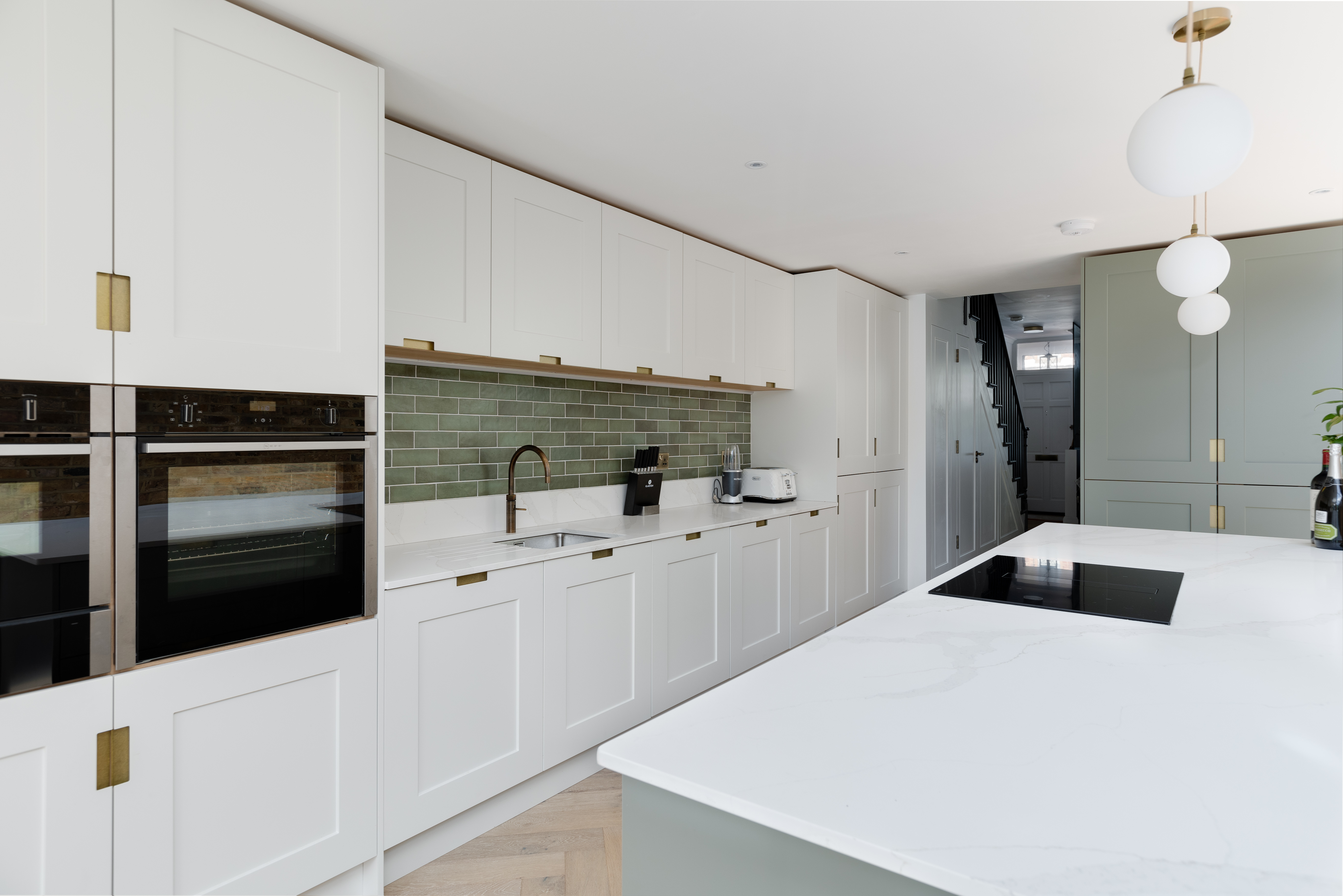 Modern kitchen featuring sleek white cabinetry, a green tiled backsplash, and a spacious island with an integrated hob. An Arch KBB team member is present, showcasing contemporary design elements and functionality in a bright, airy space.