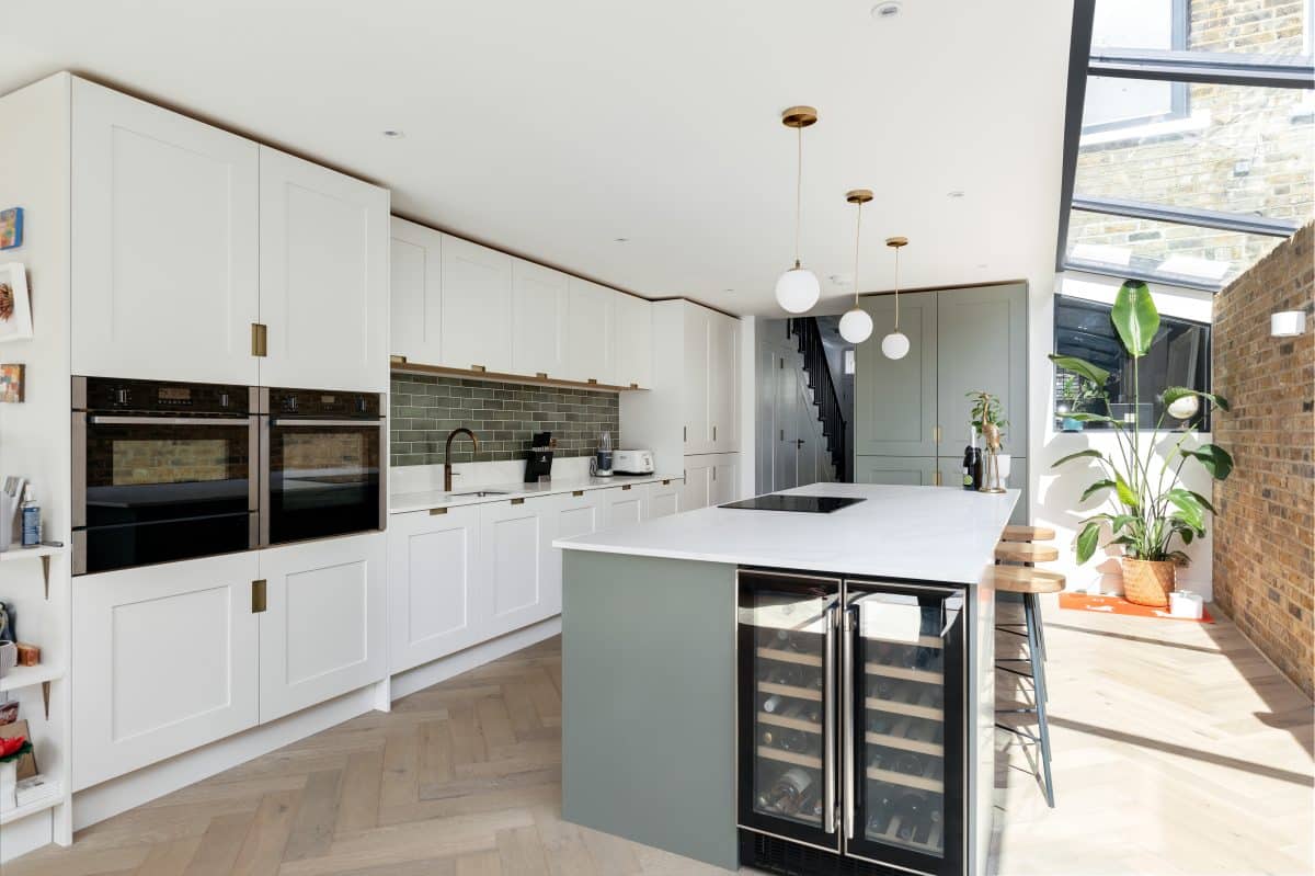 Modern kitchen interior by Arch KBB featuring sleek white cabinetry, integrated appliances, a spacious island with wine cooler, pendant lighting, and large windows allowing natural light.