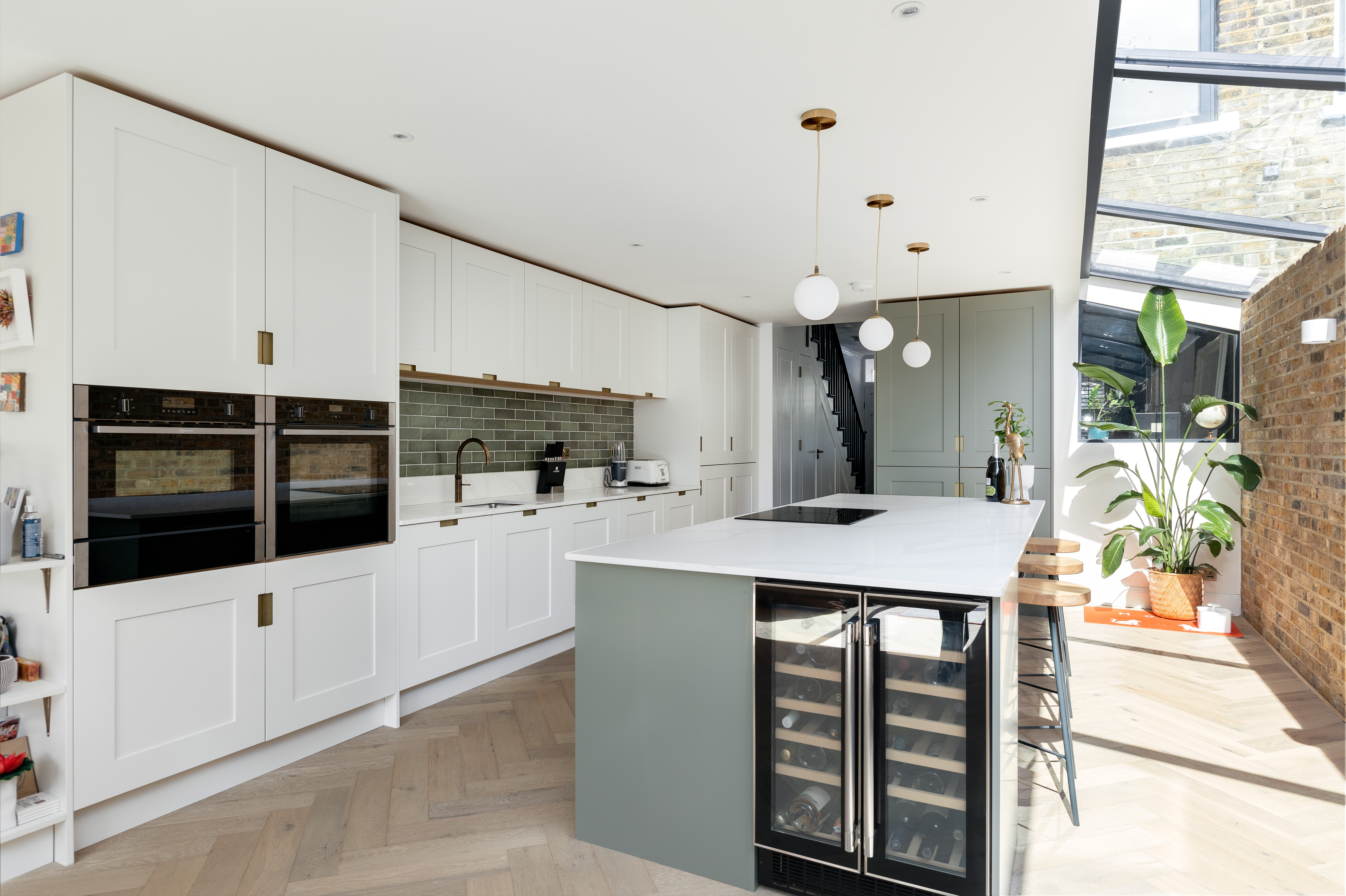 Modern kitchen design featuring a spacious island with a wine cooler, sleek cabinetry, and stylish pendant lighting. The room is illuminated by natural light from large windows, complemented by green tile backsplash and herringbone flooring. An Arch KBB team member is present, showcasing the contemporary aesthetic.