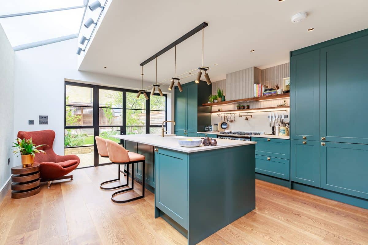 Modern kitchen design featuring a spacious island with a light-coloured countertop, complemented by teal cabinetry and stylish pendant lights. An Arch KBB team member is present, showcasing the kitchen's functional layout. Large windows provide natural light and a view of the garden, enhancing the inviting atmosphere.