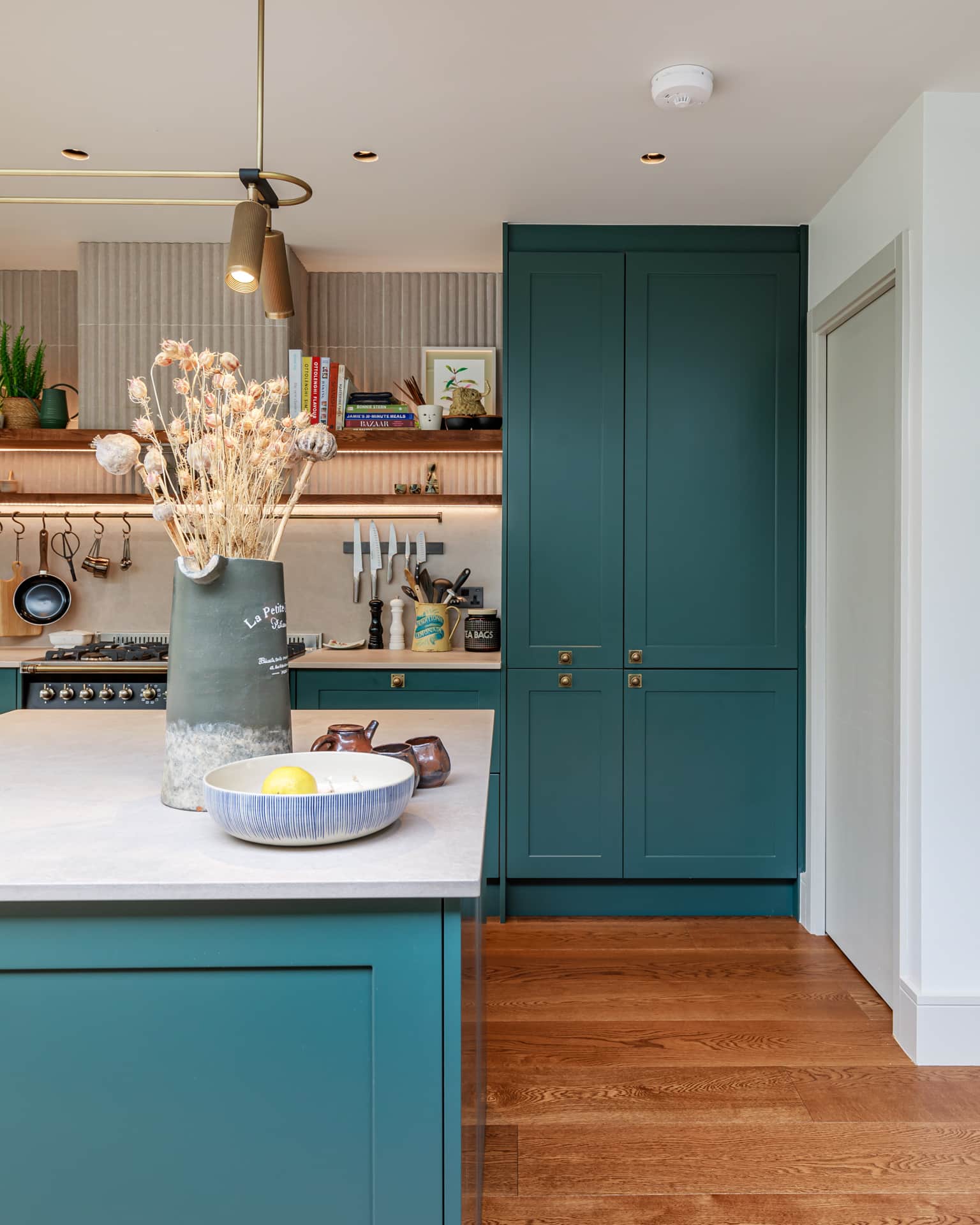 Stylish kitchen featuring a central island with a bowl of fruit, a decorative vase with dried flowers, and modern cabinetry in a rich green hue. The background includes a well-equipped cooking area with utensils and shelves filled with books and decorative items, creating a warm and inviting atmosphere. Arch KBB team member present, contributing to the design expertise.