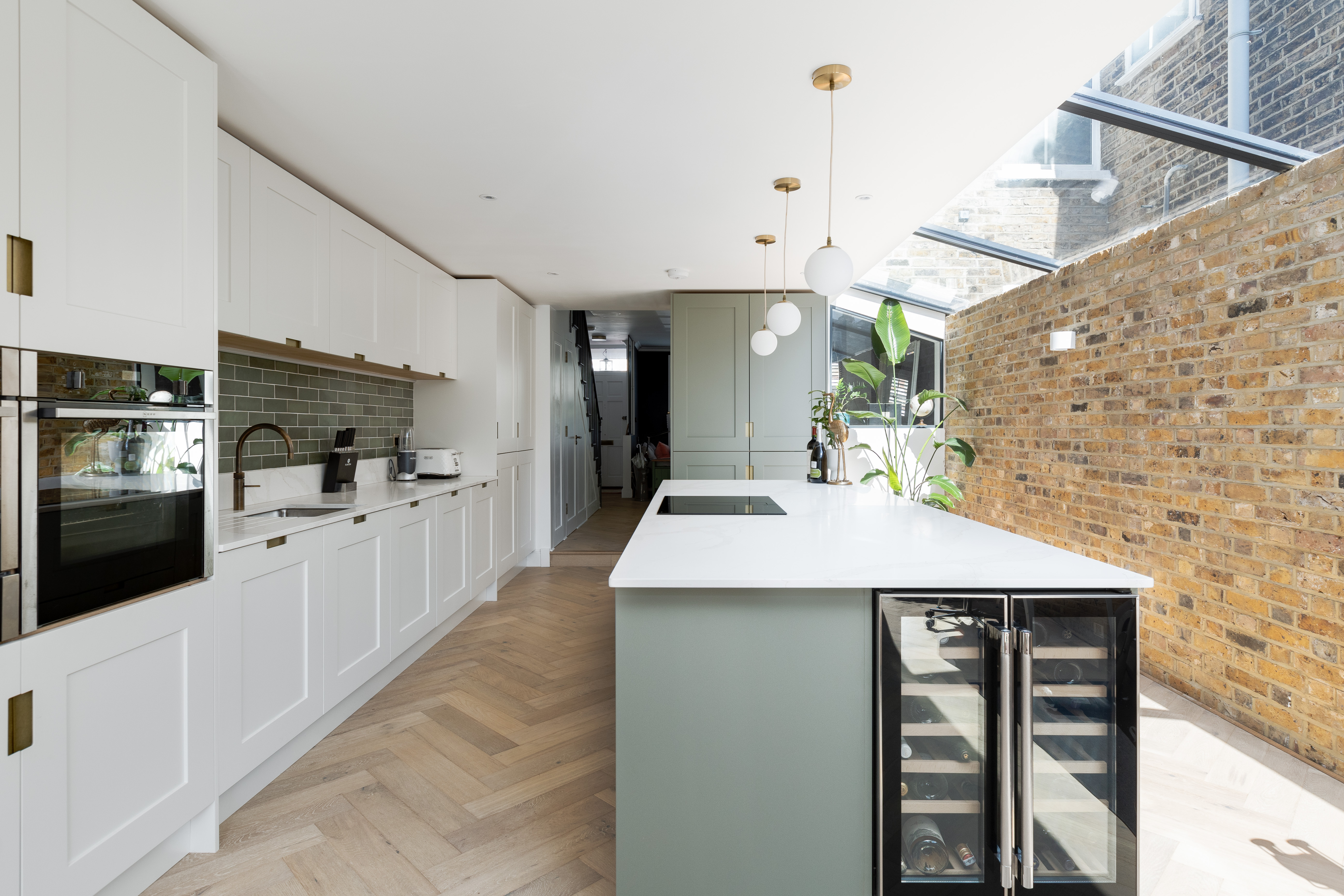 Modern kitchen design featuring a spacious layout, elegant cabinetry, and stylish pendant lighting. The open area includes a central island with a cooktop, integrated appliances, and a wine cooler, complemented by natural light from a skylight. An Arch KBB team member is present, enhancing the design experience.