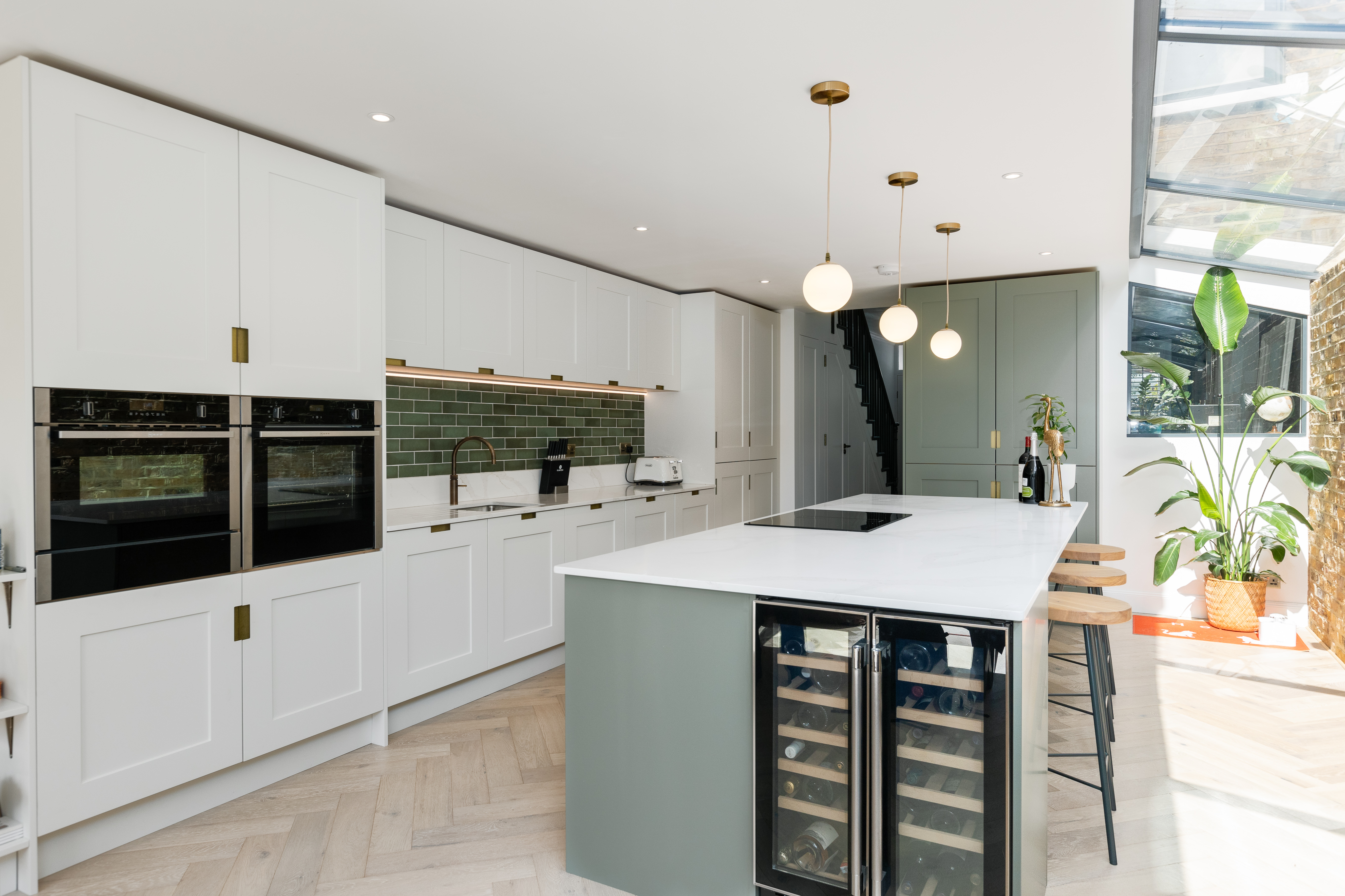 Modern kitchen design featuring a spacious island with a wine cooler, sleek cabinetry in white and green, and stylish pendant lighting. An Arch KBB team member is present, showcasing the contemporary layout and elegant finishes, including a herringbone floor and green tiled backsplash. Natural light floods the space through large windows, enhancing the inviting atmosphere.