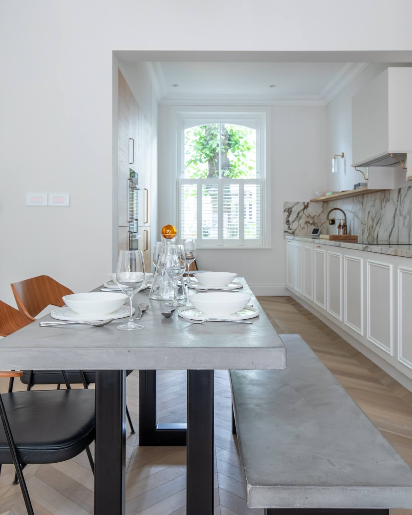 Modern kitchen interior featuring a concrete dining table set for a meal, complete with white plates and glassware. The space is bright and airy, highlighted by natural light from a large window with greenery outside. Sleek cabinetry and marble countertops complement the contemporary design, creating an inviting atmosphere for dining. An Arch KBB team member is present, contributing to the scene.