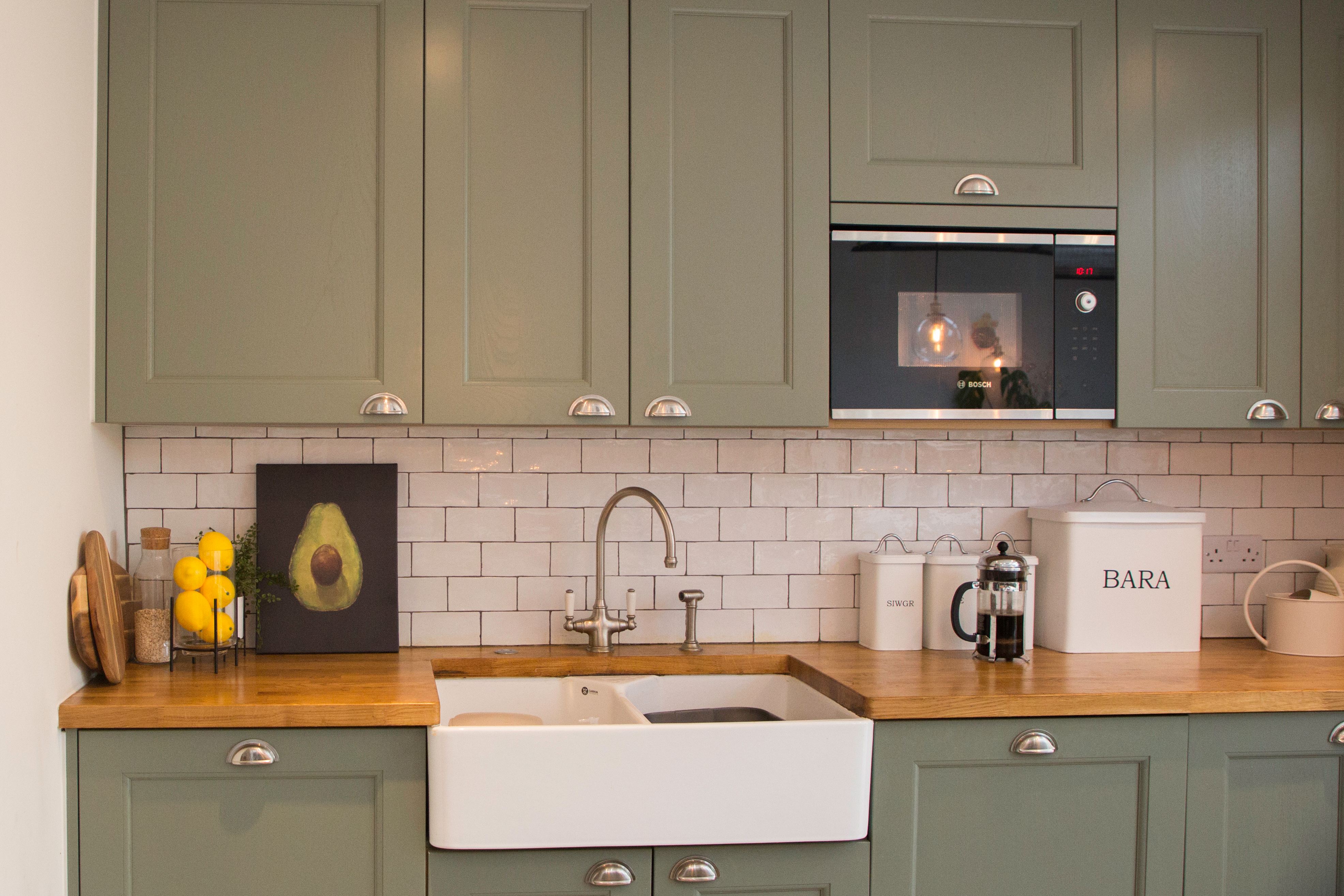 Modern kitchen featuring green cabinetry, a wooden countertop, and a farmhouse sink. The backsplash is tiled with white subway tiles, adorned with a framed avocado artwork and fresh lemons. An Arch KBB team member is present, showcasing a stylish microwave and kitchen accessories.