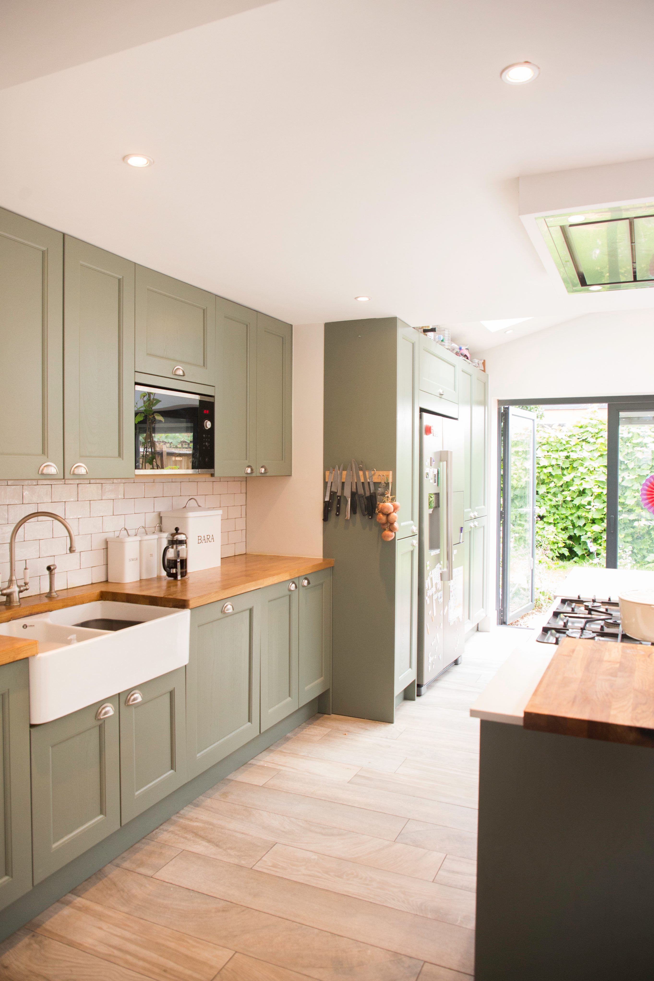 Modern kitchen featuring soft green cabinetry, a farmhouse sink, and wooden countertops. The space includes a built-in microwave, a stainless steel fridge, and an open door leading to an outdoor area. An Arch KBB team member can be seen, contributing to the design aesthetic.
