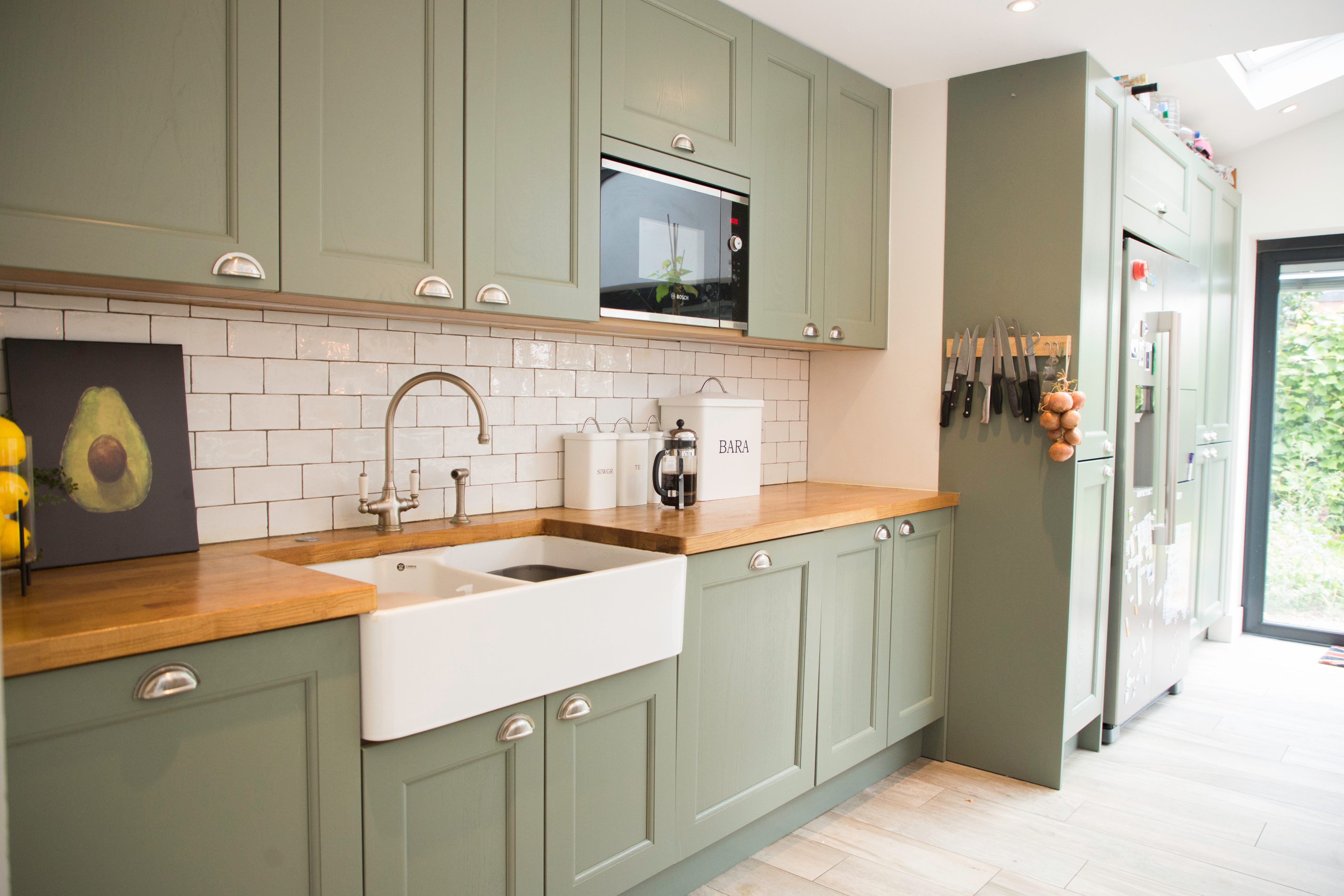 Modern kitchen featuring green cabinetry, a farmhouse sink, and wooden countertops. The space includes a microwave and stylish backsplash tiles, with kitchen utensils neatly arranged. An Arch KBB team member is present, contributing to the kitchen design.