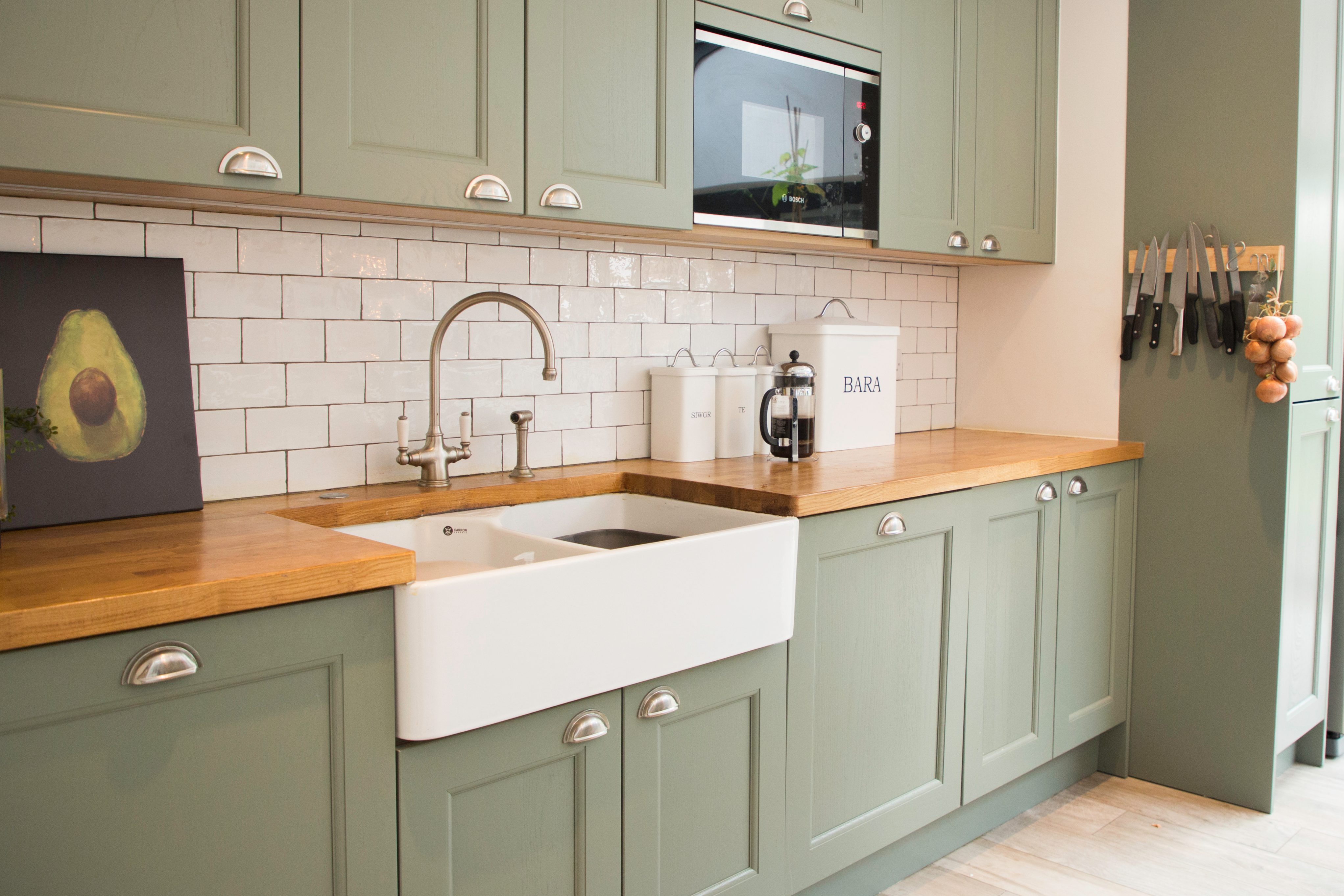 Modern kitchen featuring a farmhouse sink, green cabinetry, and a wooden countertop. The design includes white tiled backsplash, a stylish tap, and kitchen essentials displayed on the counter. An Arch KBB team member can be seen in the background, contributing to the kitchen's design.