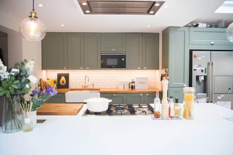 Modern kitchen design featuring green cabinetry, a white countertop, and a gas stove. The space includes a stylish sink, built-in microwave, and various kitchen appliances. Decorative elements include a vase with flowers and a bowl of pasta. An Arch KBB team member is present, contributing to the kitchen's inviting atmosphere.